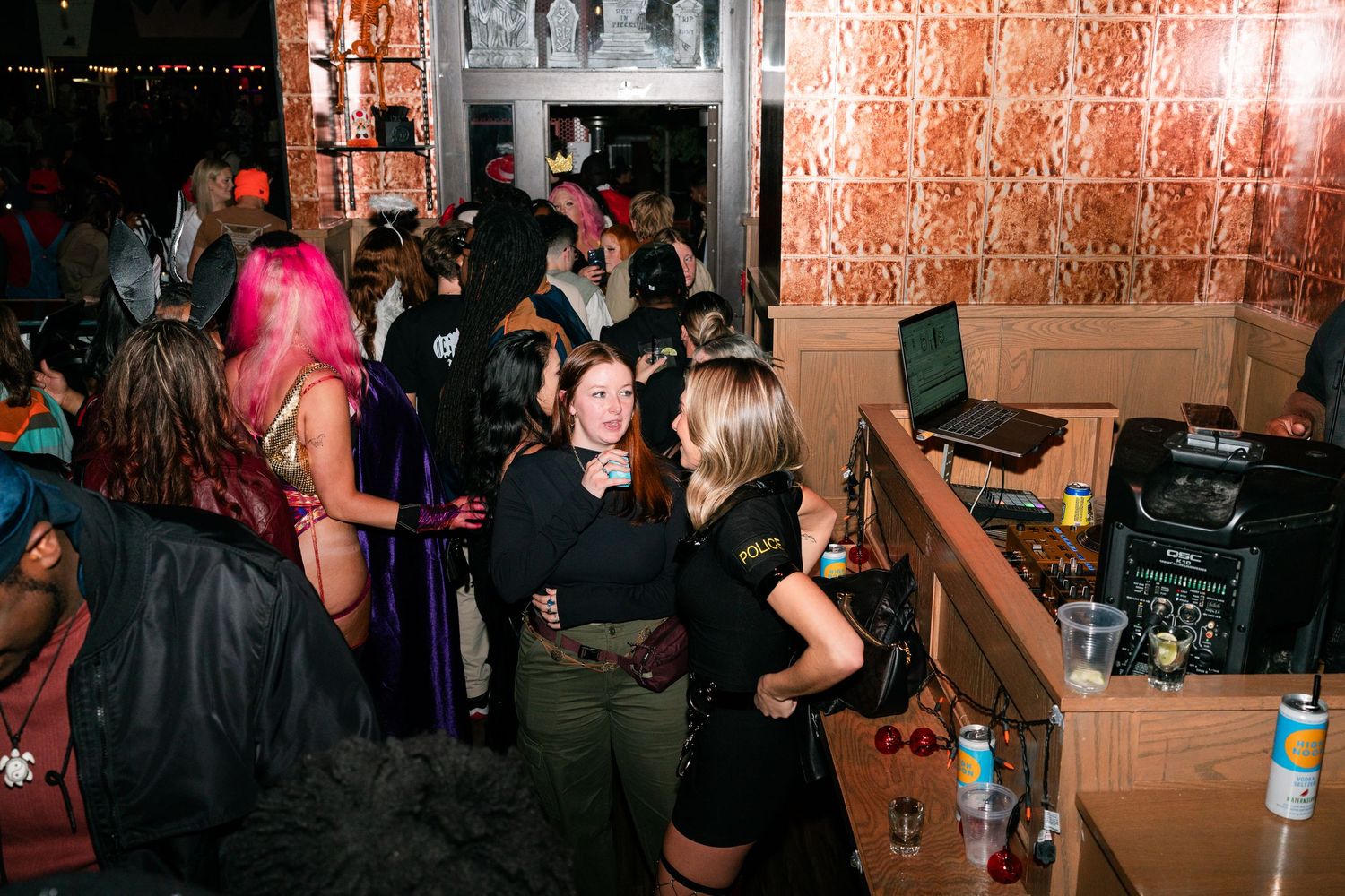 Women in Halloween costumes socializing near a DJ booth at a packed bar.