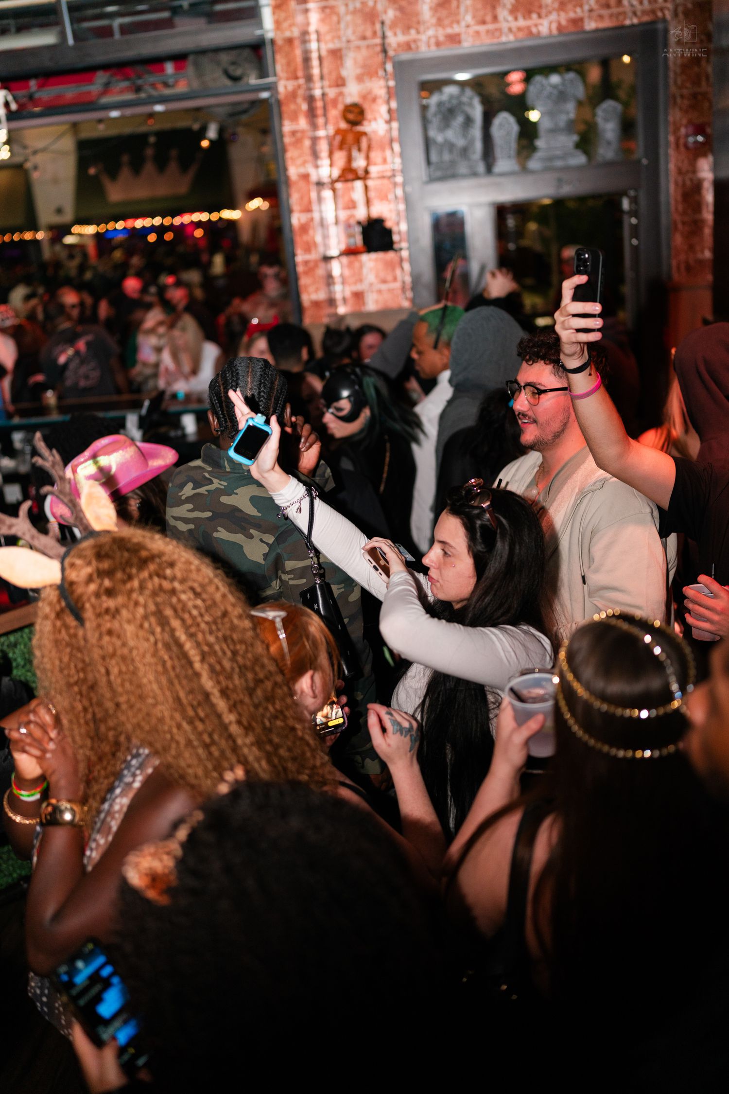Diverse crowd at Halloween party with people holding phones and drinks in decorated venue.