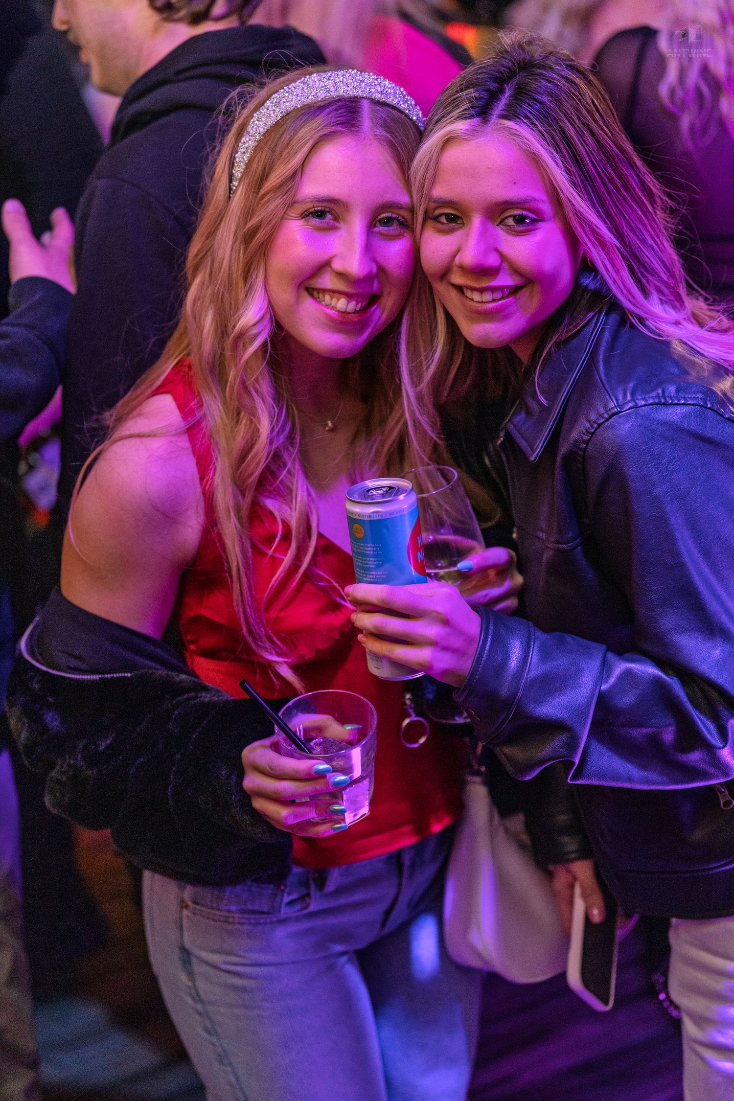 Two young women smile holding drinks under pink and purple lights at an indoor party.