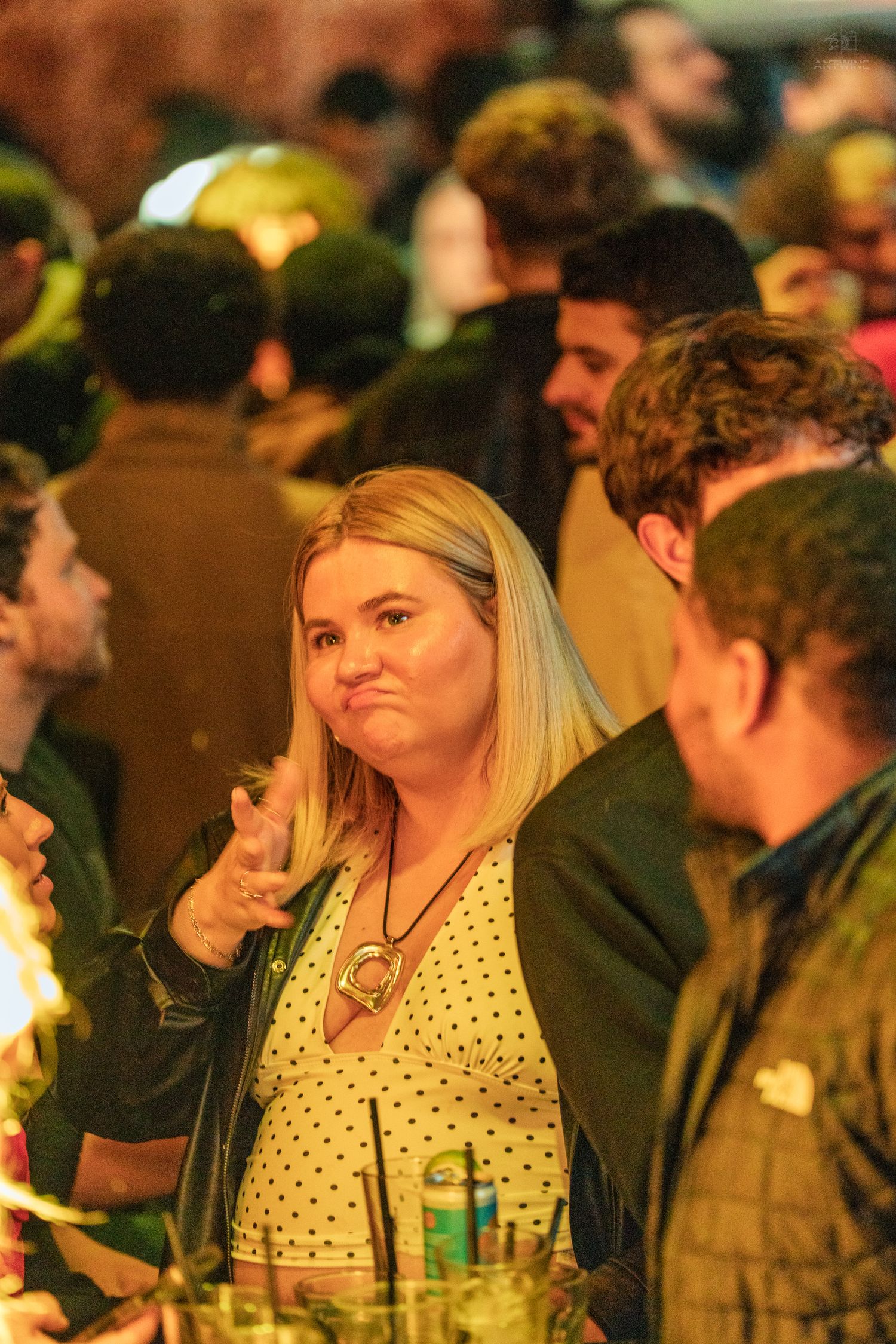 Blonde woman in polka dot top making an expressive face at a busy social event.