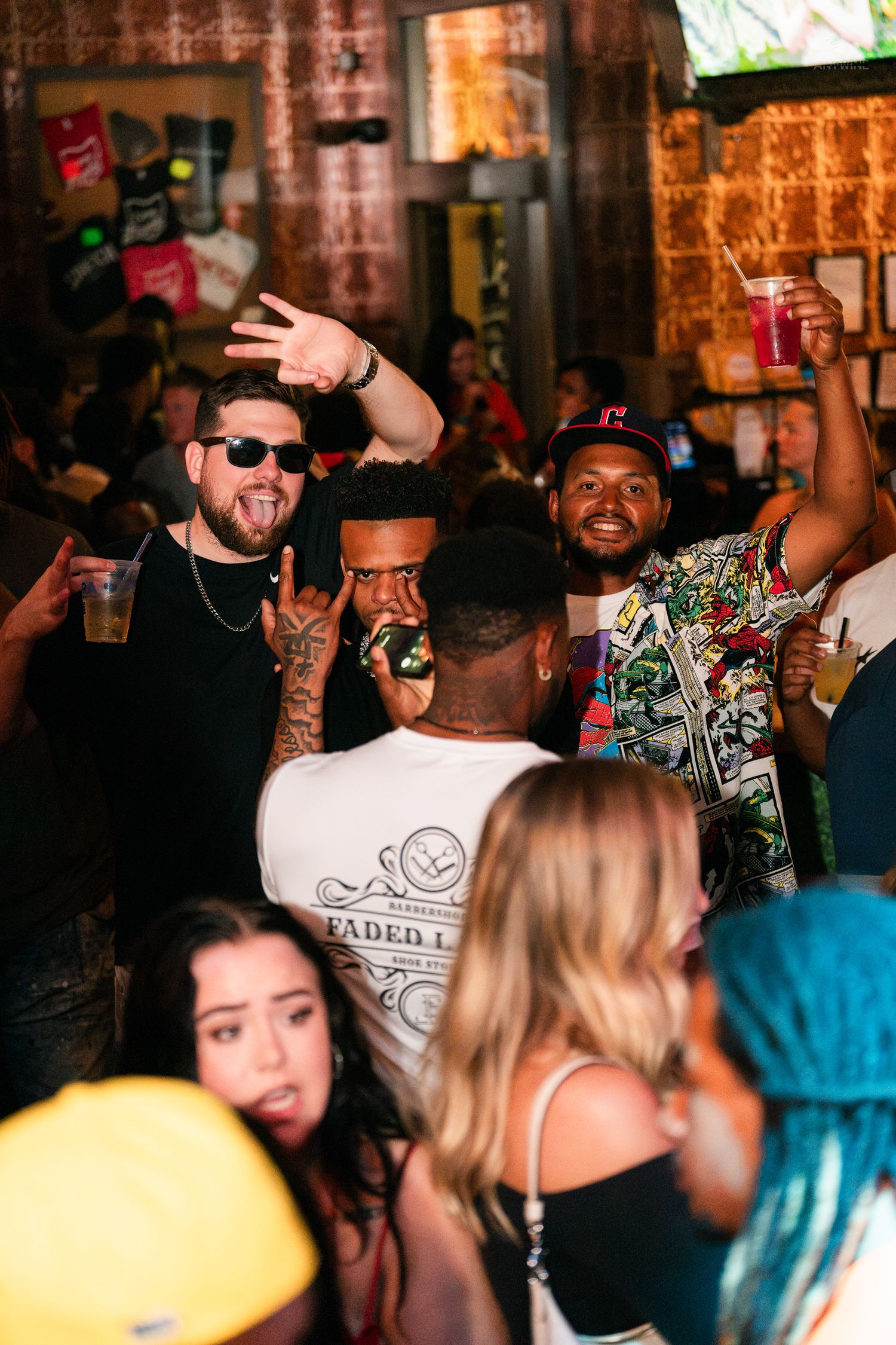 Lively bar crowd with tattooed man making rock sign, friends holding drinks and celebrating at a nightlife event.