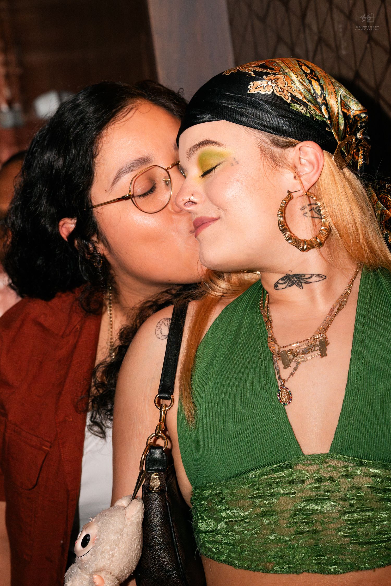 Two women sharing a kiss at a party, one wearing green eyeshadow and gold hoop earrings.