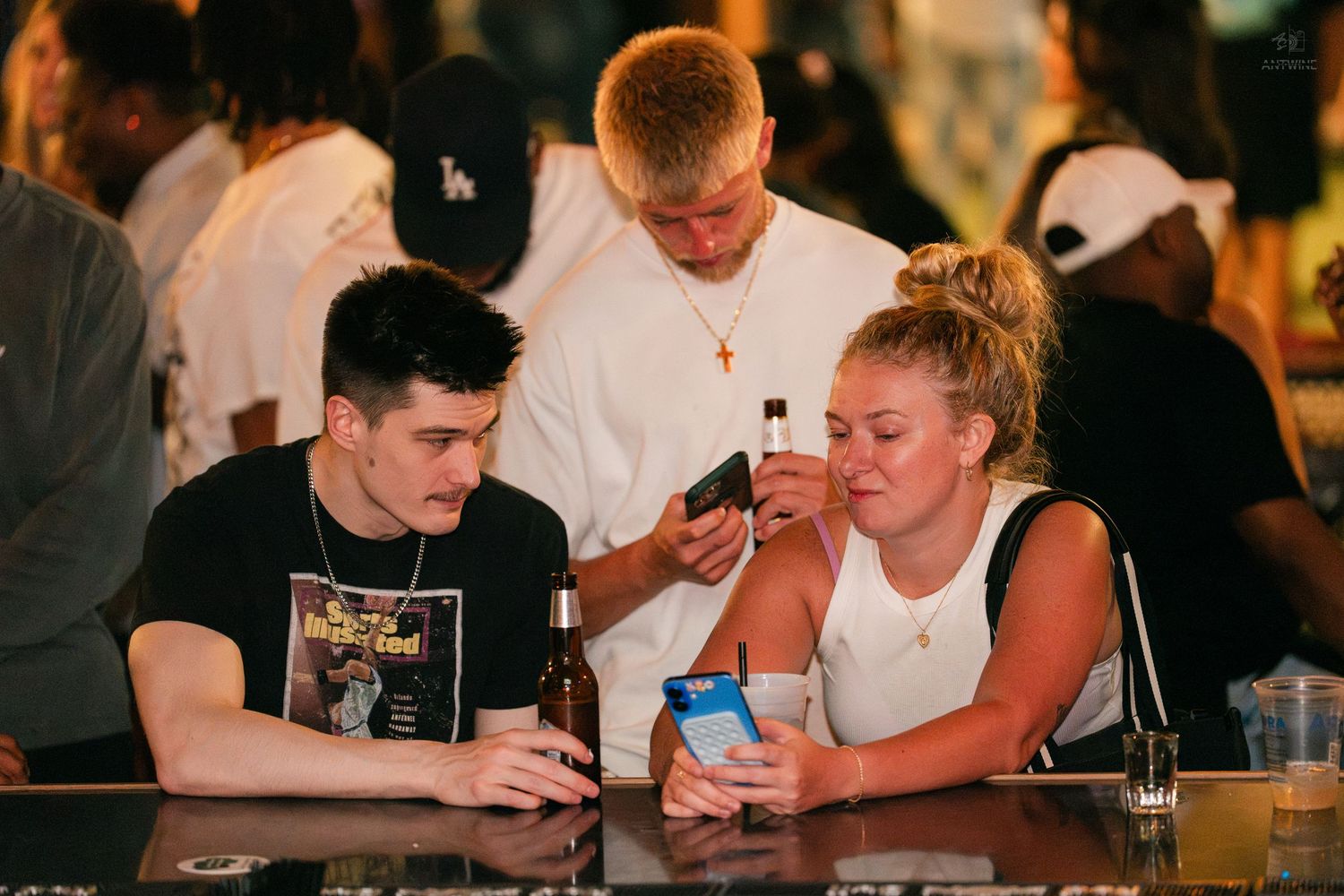 A young couple looks at a phone together at a crowded bar, drinks in hand.