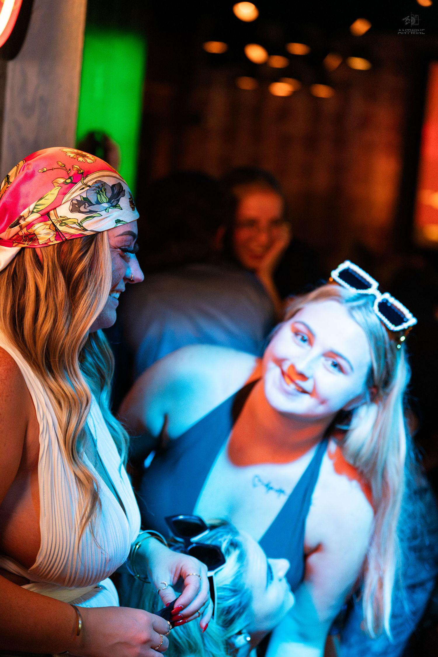 Two blonde women chatting and smiling under colorful lights at a nightclub.