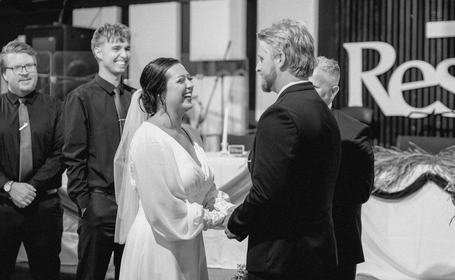 Black and white wedding photo of bride and groom holding hands, smiling, with guests in background.