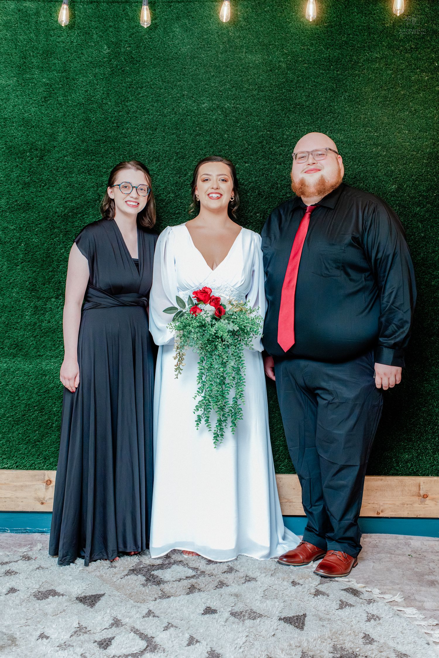 Bride in white gown holding red bouquet poses with bridesmaid in black dress and groomsman in black suit before green wall.