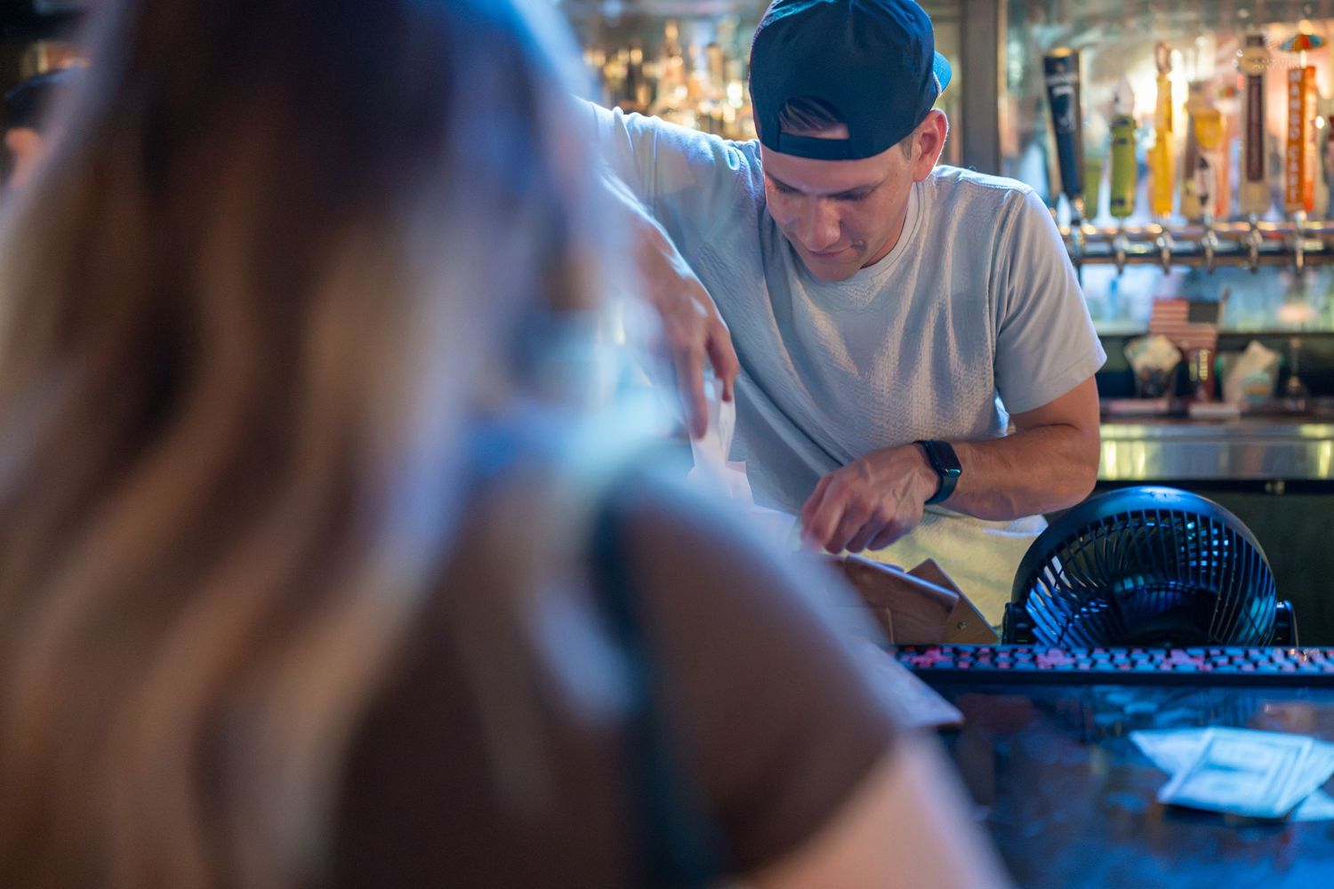 Bartender in a cap reviews menu with a customer at a busy bar counter with draft beer taps in background.