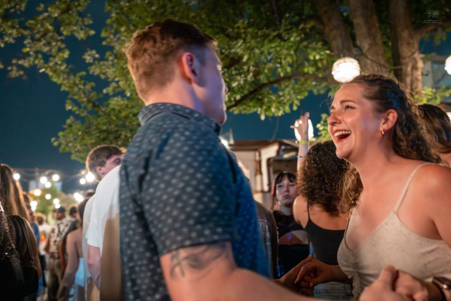 A couple laughs and dances together at an outdoor nighttime festival event with string lights and trees.