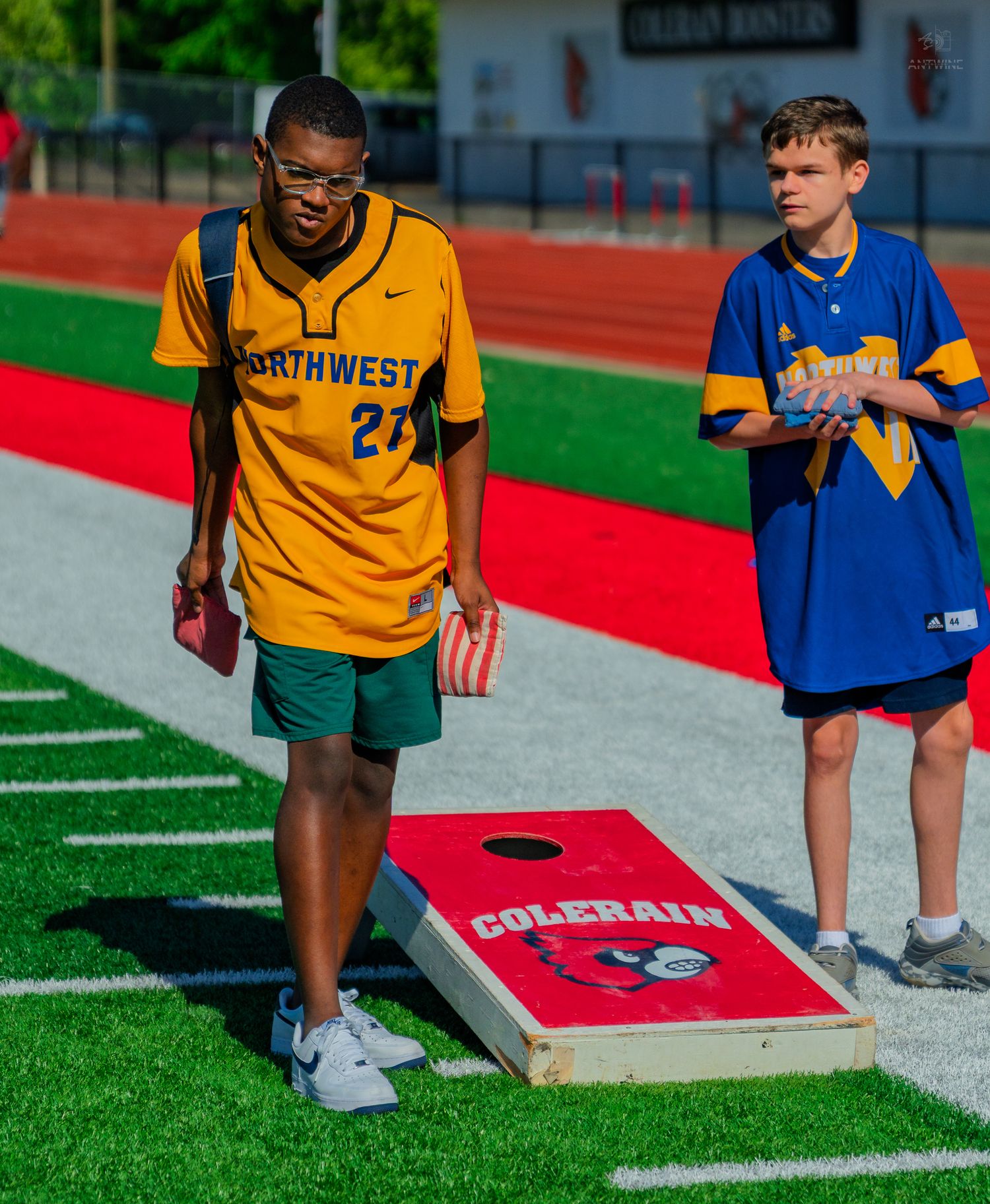 Two boys playing cornhole on a red turf field, one wearing a Northwest jersey and the other in a blue Colerain uniform.