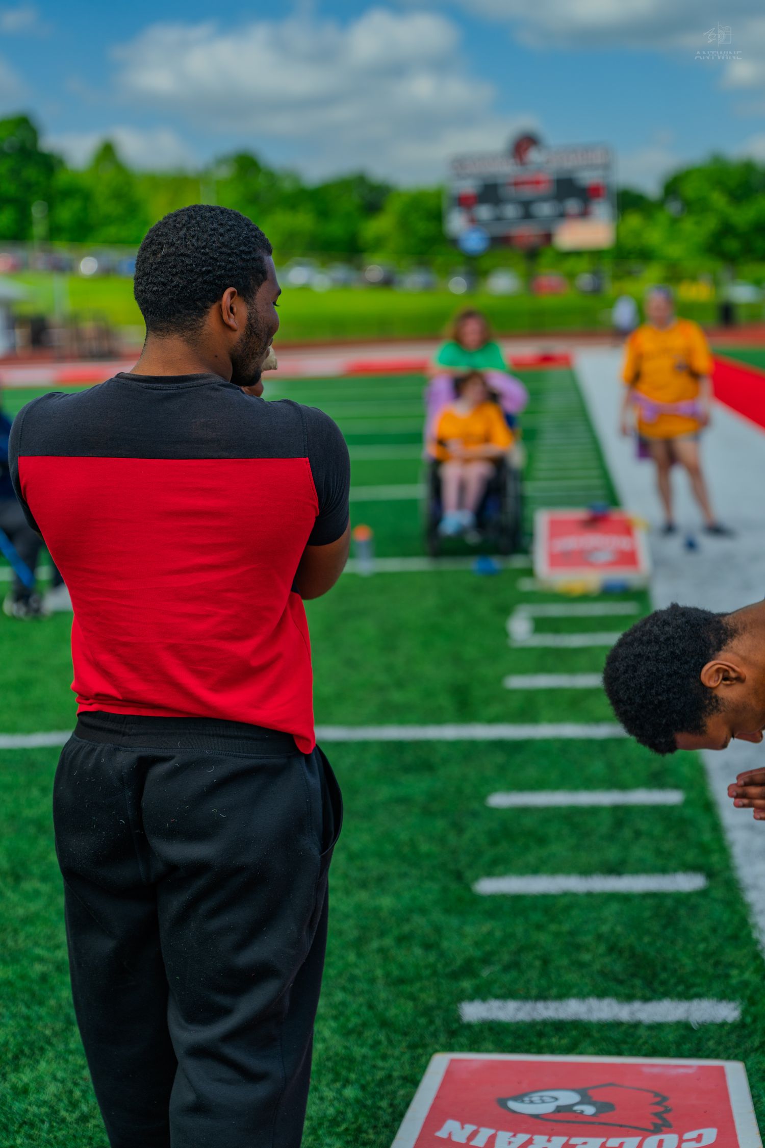 Athletes and coaches gather on a red track field during an outdoor sports event on a sunny day.