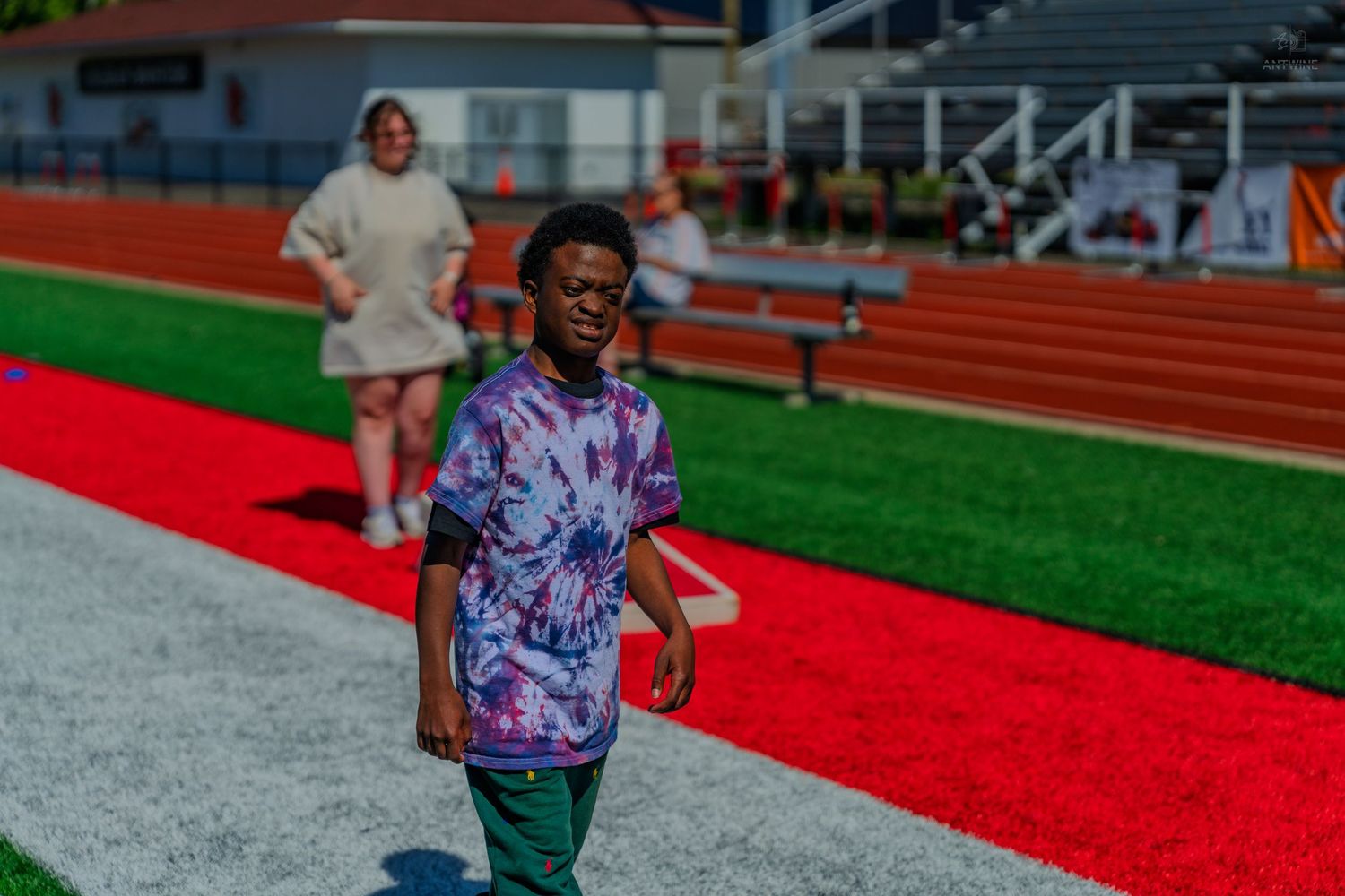A young boy in a tie-dye shirt walks on a red track at a stadium, with people and bleachers in the background.