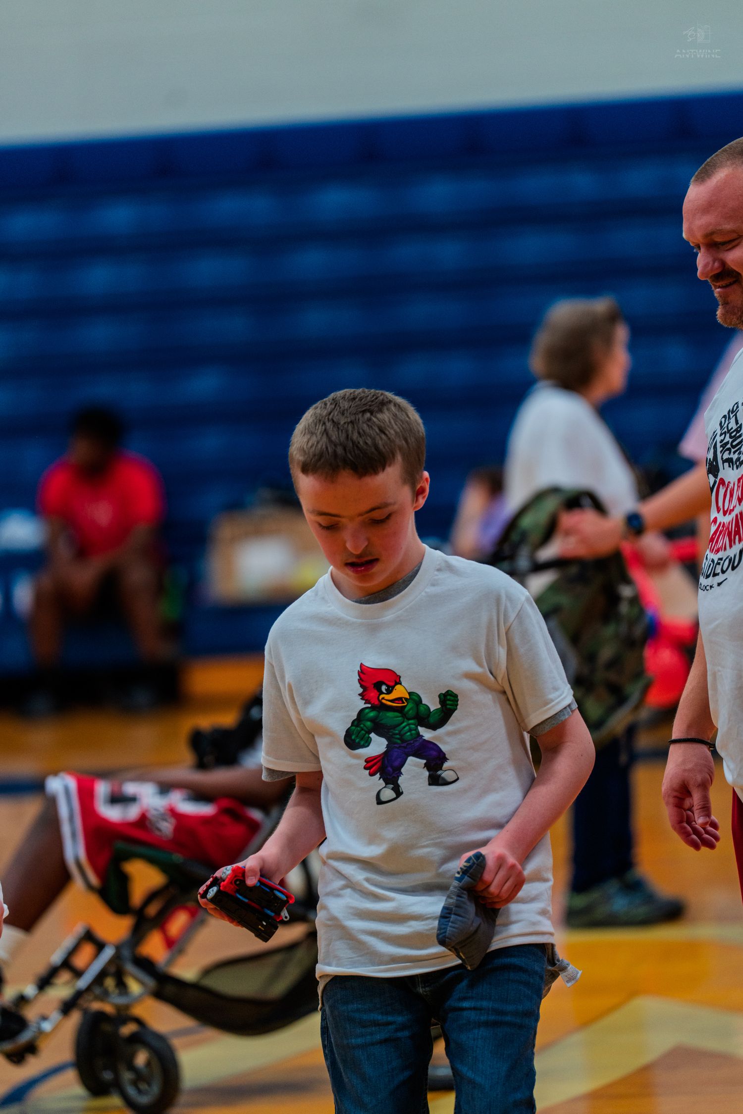 Boy in cardinal mascot t-shirt walks in gymnasium during school event with adults and children in background.