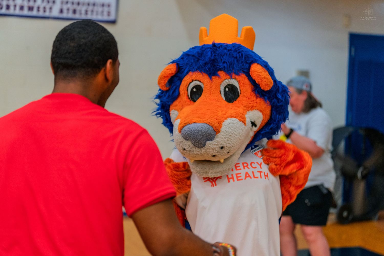 Mercy Health mascot in orange and blue costume interacting with attendees at an indoor sports event.