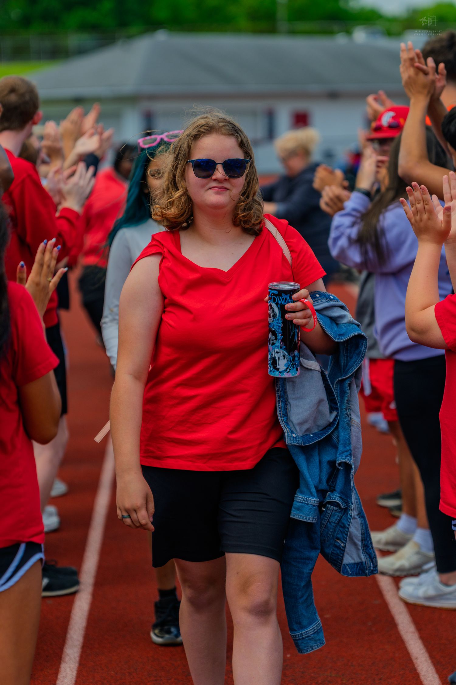 Young woman in red shirt and sunglasses walks through cheering crowd on a track during a school event.