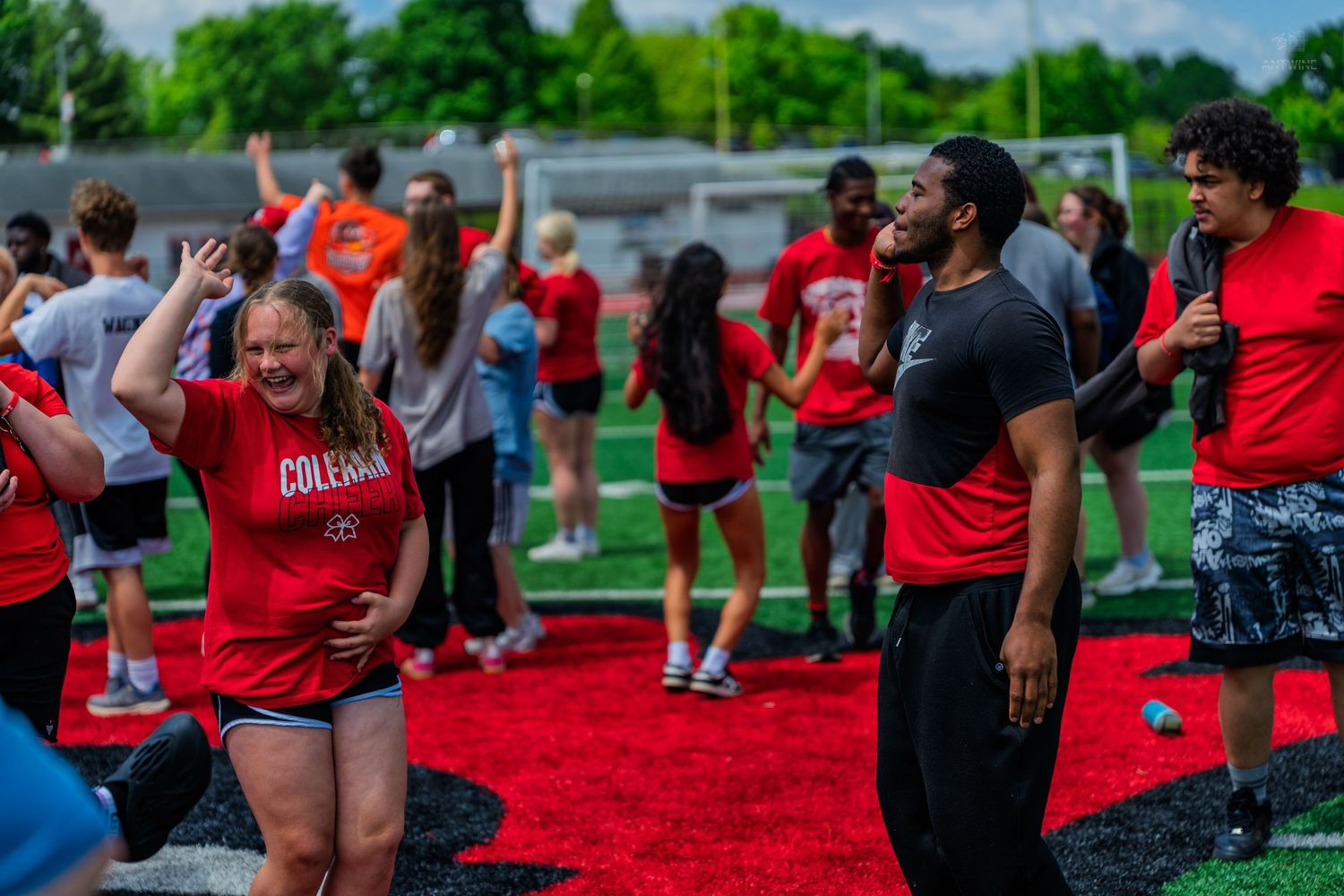 Students in red shirts celebrate outdoors on a turf field during a college event on a sunny day.