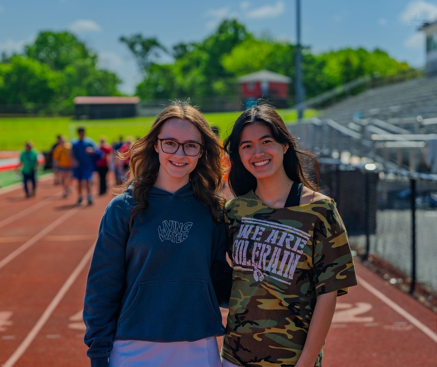 Two teenage girls smiling together on a school track field on a sunny day, one wearing a navy hoodie and the other a camo shirt.