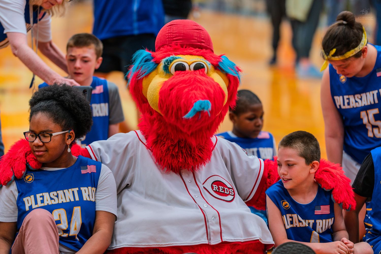 Cincinnati Reds mascot Mr. Redlegs poses with youth basketball players in Pleasant Run uniforms at a gym event.