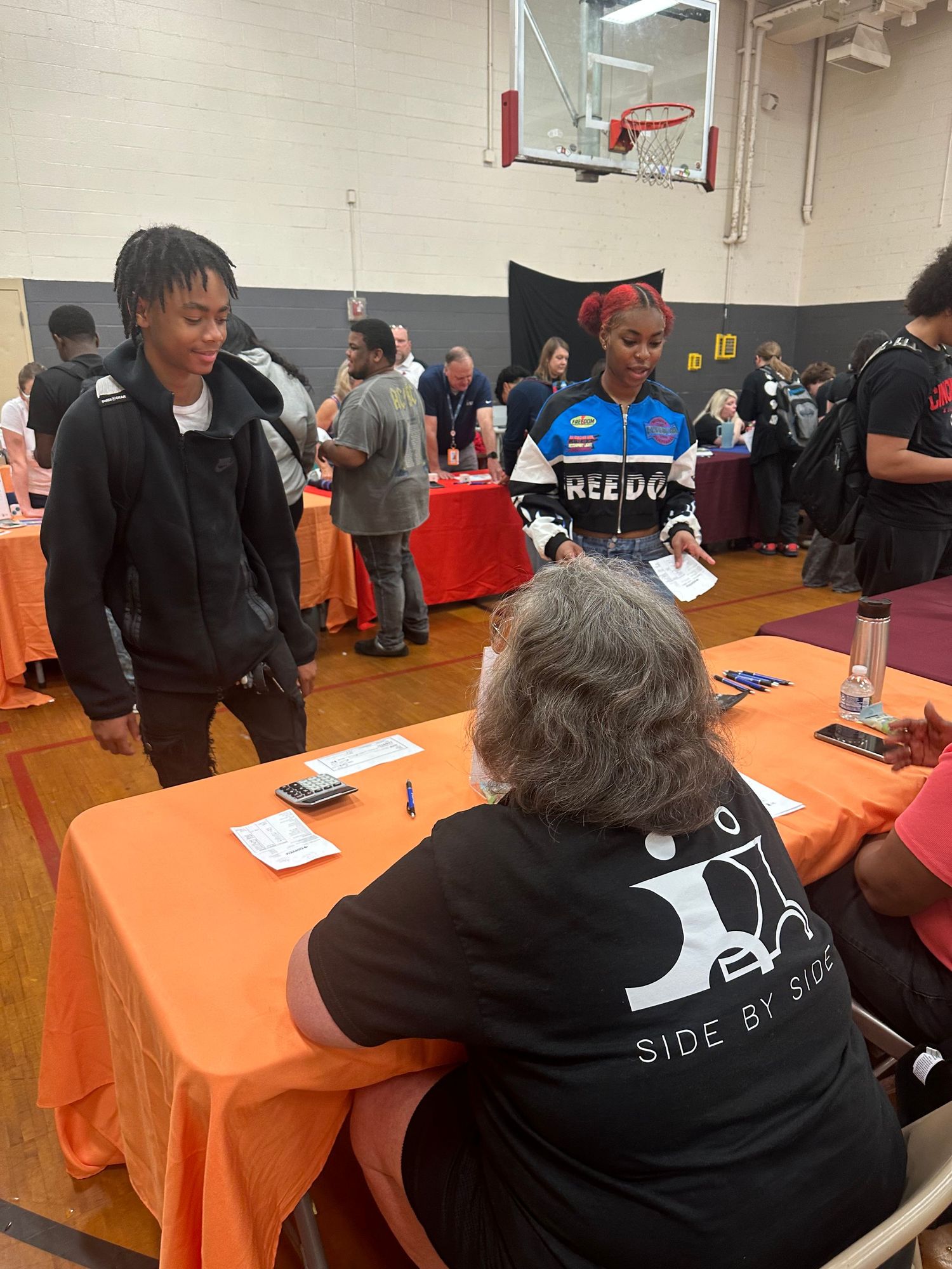 People networking at an indoor job fair in a gymnasium with orange tablecloths and vendor booths in the background.