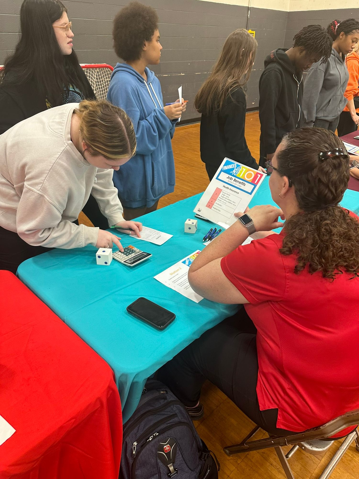 Students collaborate at a teal table during a school fair activity in a gymnasium, using calculators and worksheets.