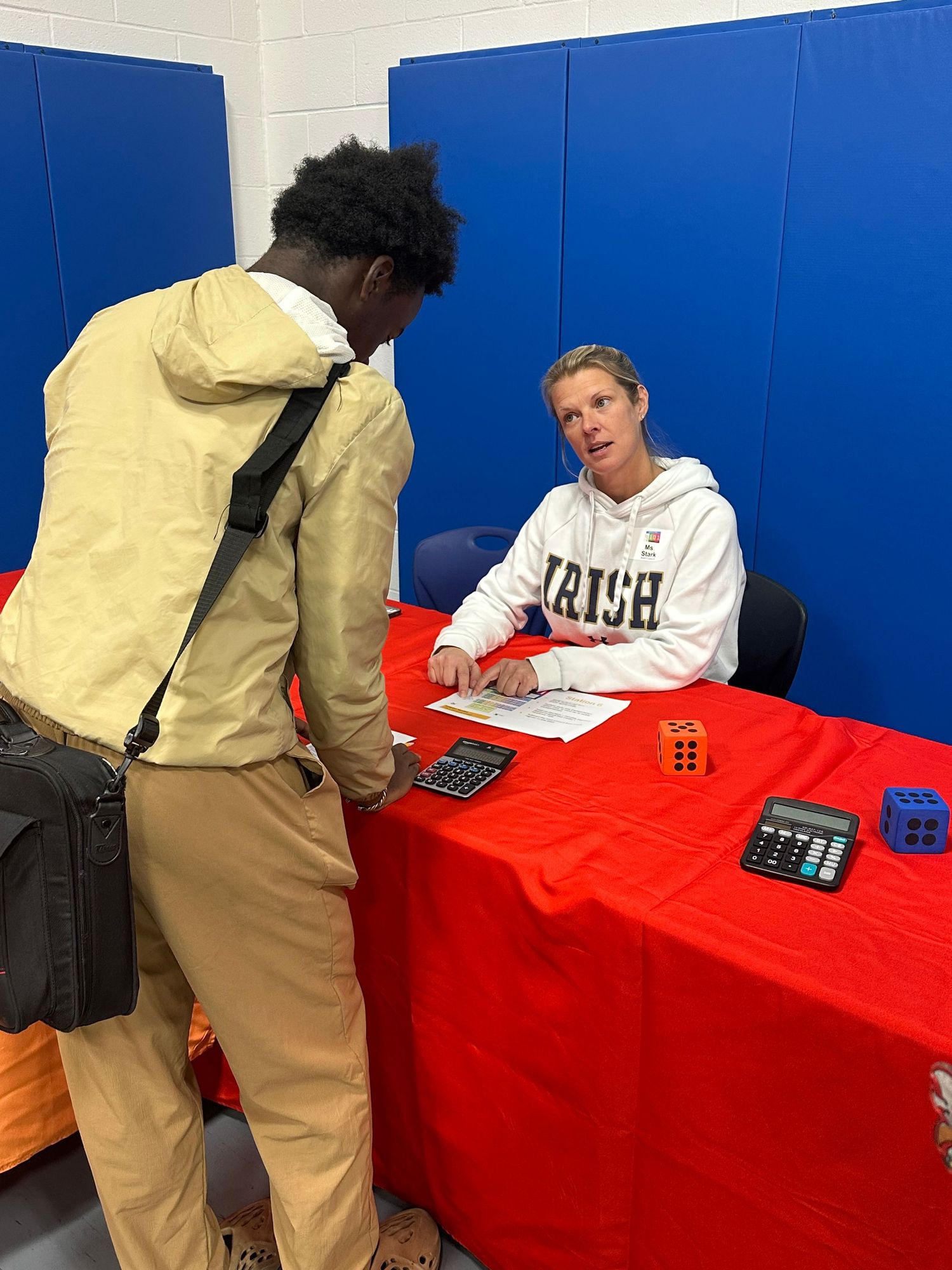 A woman in an Irish hoodie sits at a red table with calculators, speaking with a student at an indoor event.