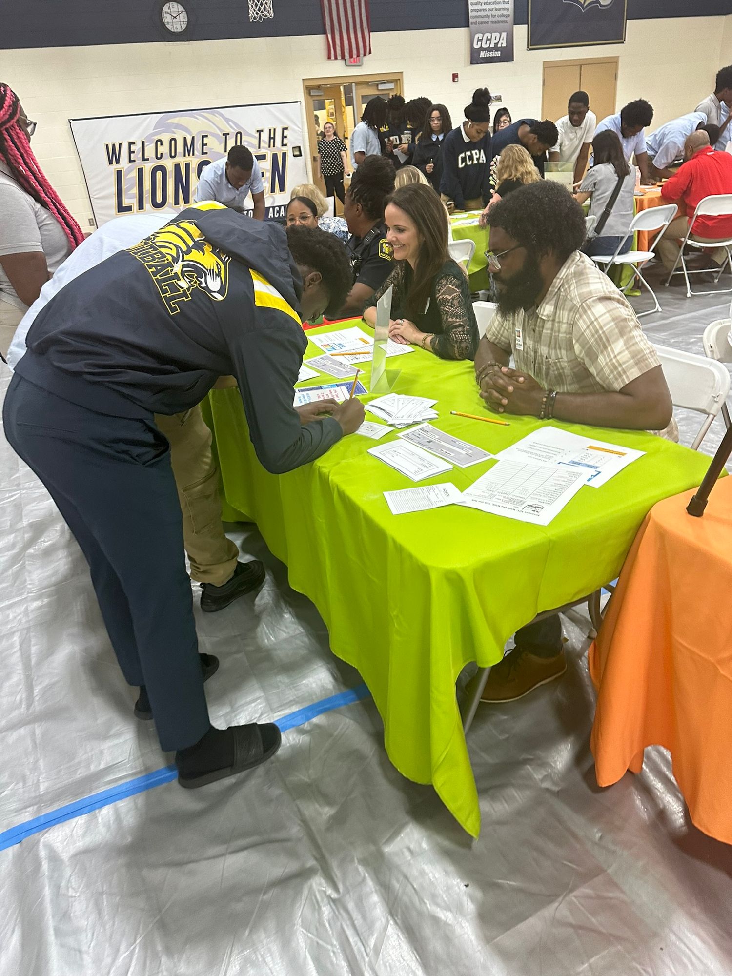 Students and staff gather at a Welcome to the Lions Den school orientation event, reviewing documents at a bright green table.