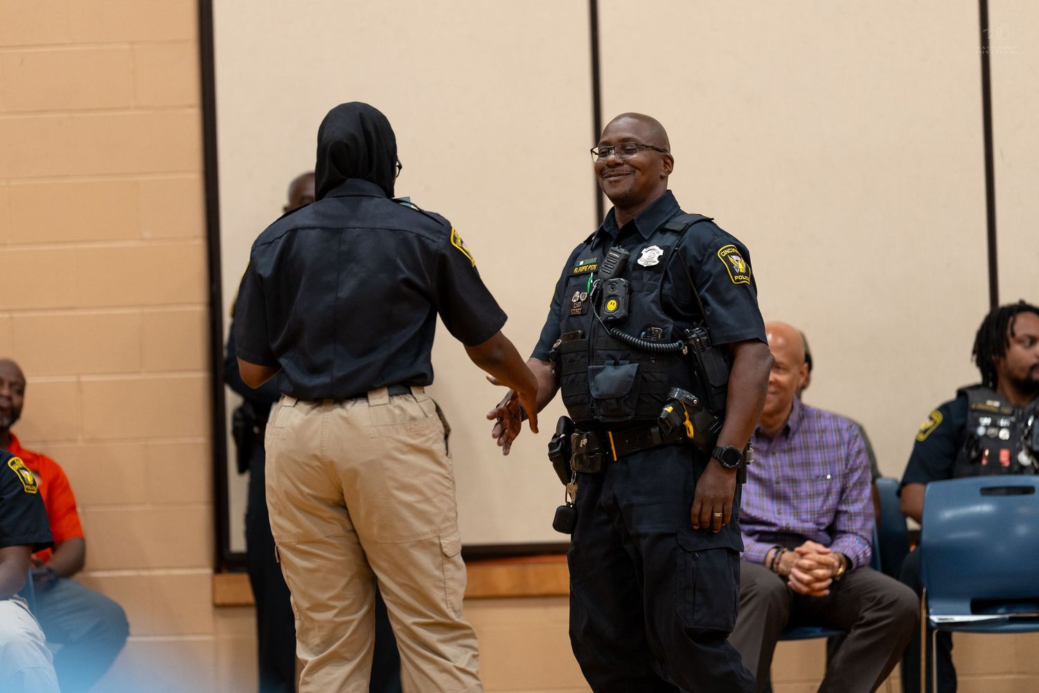 Two police officers shaking hands at a community meeting inside a public building with seated attendees in background.
