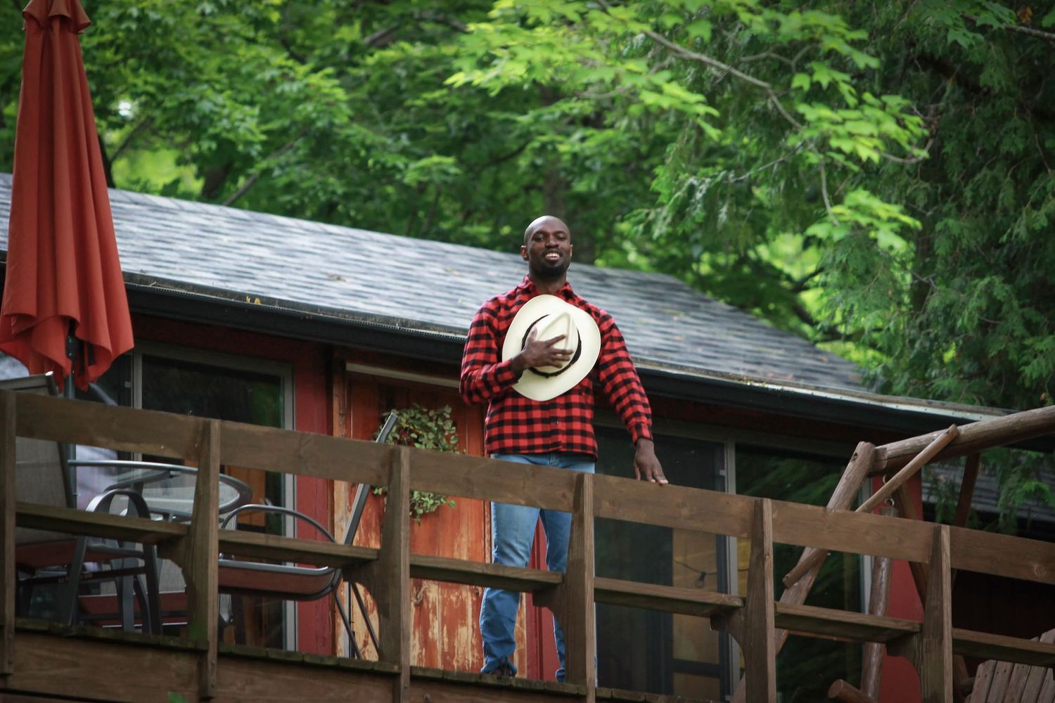 Man in plaid shirt and straw hat standing on wooden deck surrounded by green trees near a rustic cabin.