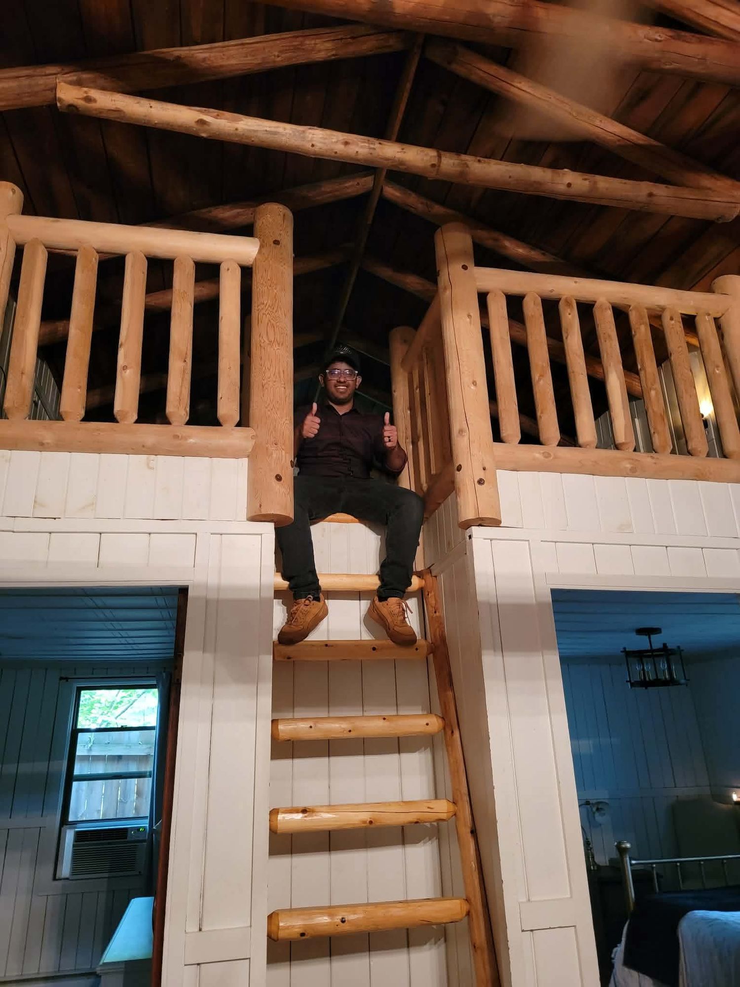 A smiling man sits on a wooden ladder leading to a rustic log cabin loft with exposed beam ceiling.