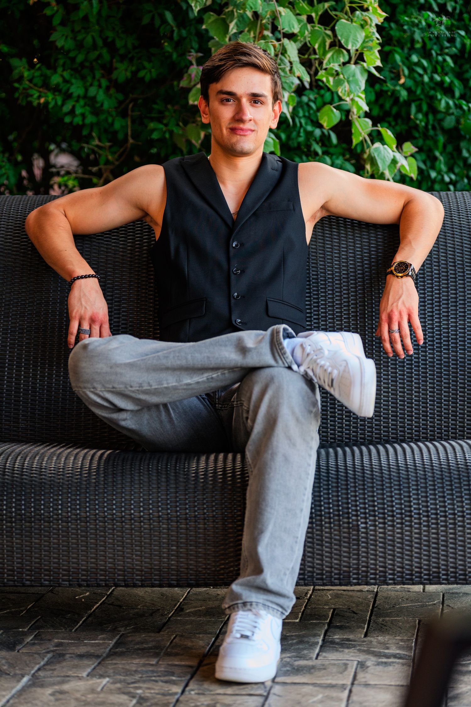 Young man in black vest and gray jeans sitting relaxed on wicker sofa outdoors.