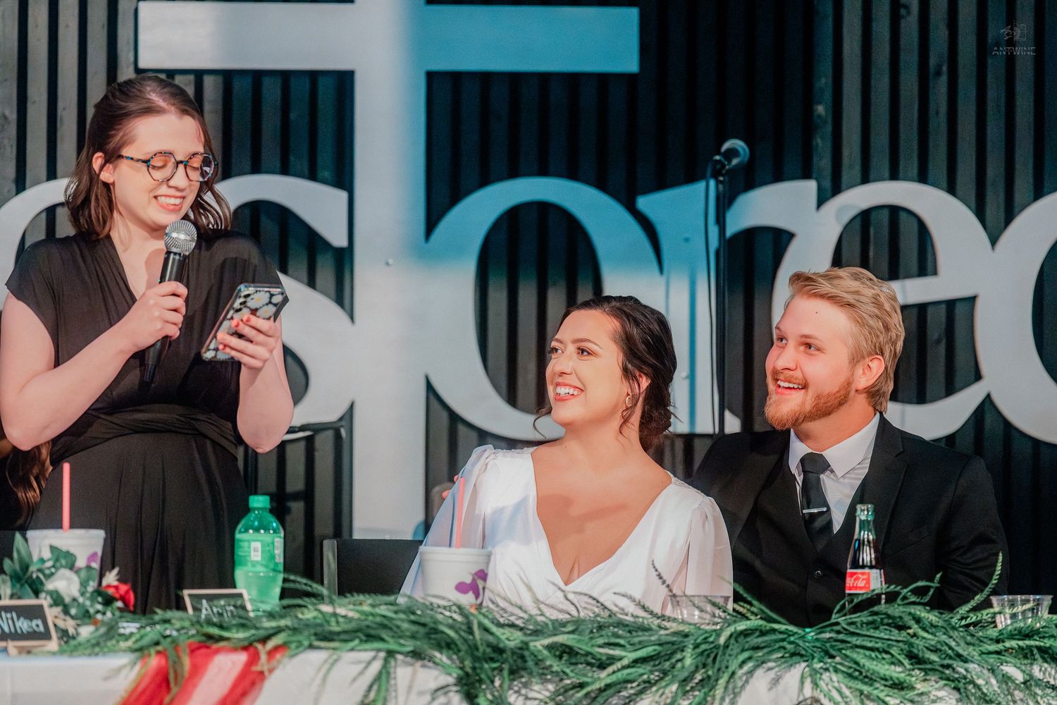 A woman speaks into a microphone giving a wedding toast as the bride and groom smile warmly.