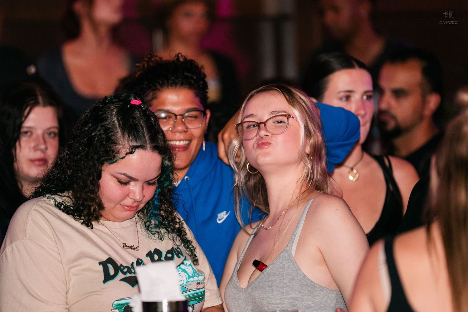 Three young women posing together in a crowded nightclub, wearing casual clothing under colorful lights.