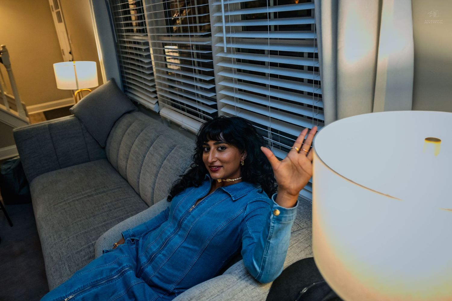 Woman in denim outfit relaxing on a couch next to a glowing lamp indoors.