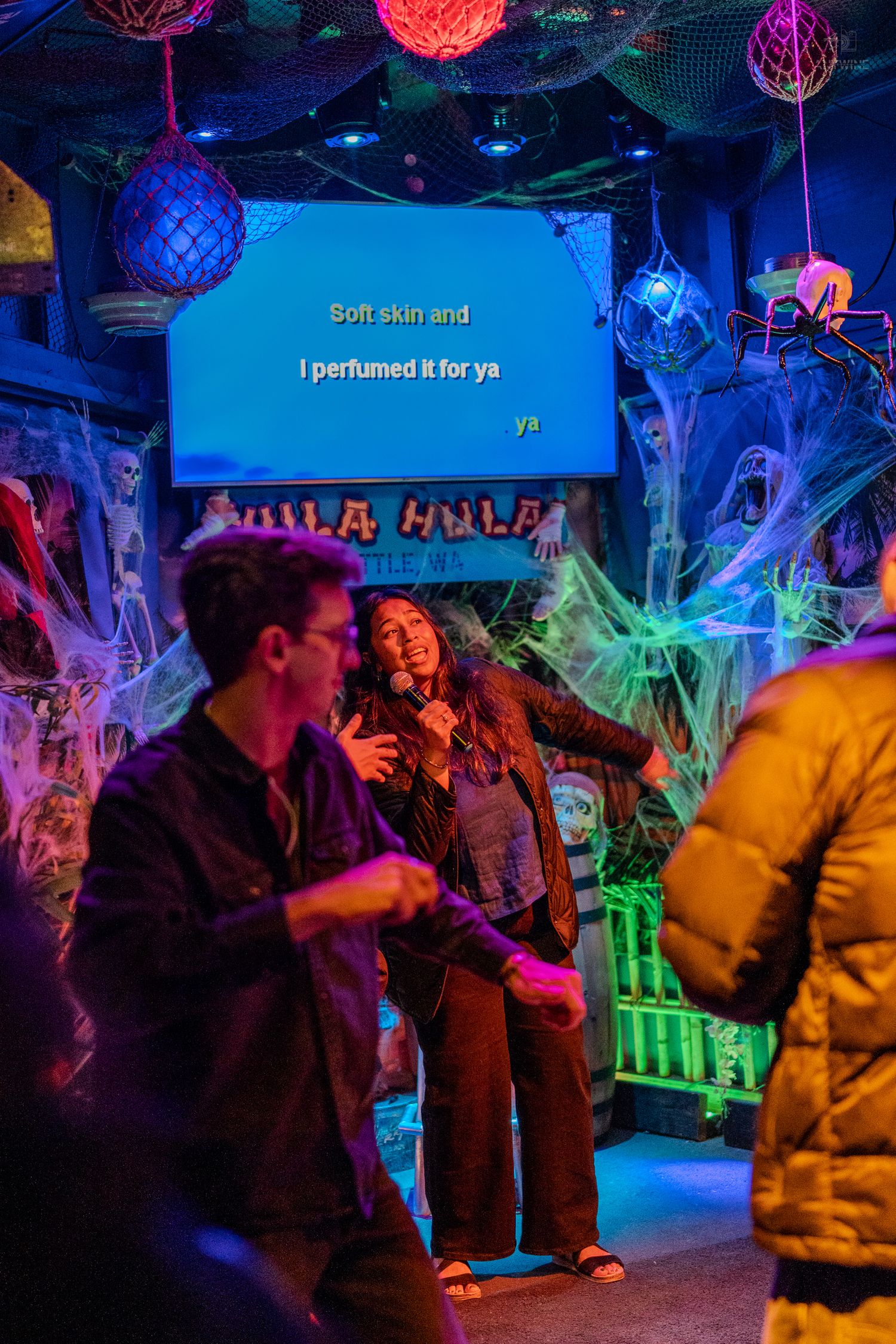 Woman performing karaoke at Hula Hula Seattle tiki bar with colorful lights and Halloween spider web decor.