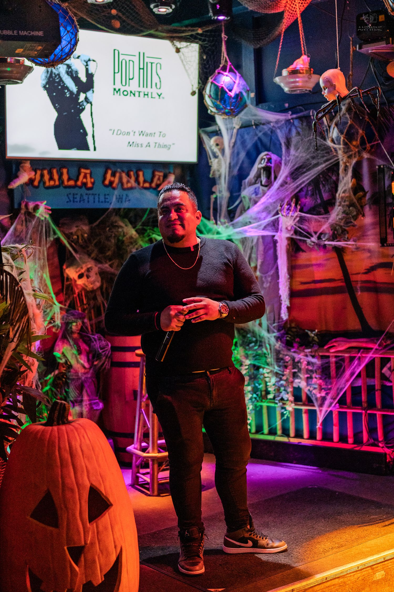Man holding microphone at Halloween karaoke bar with pumpkin and spooky decorations under neon lights.