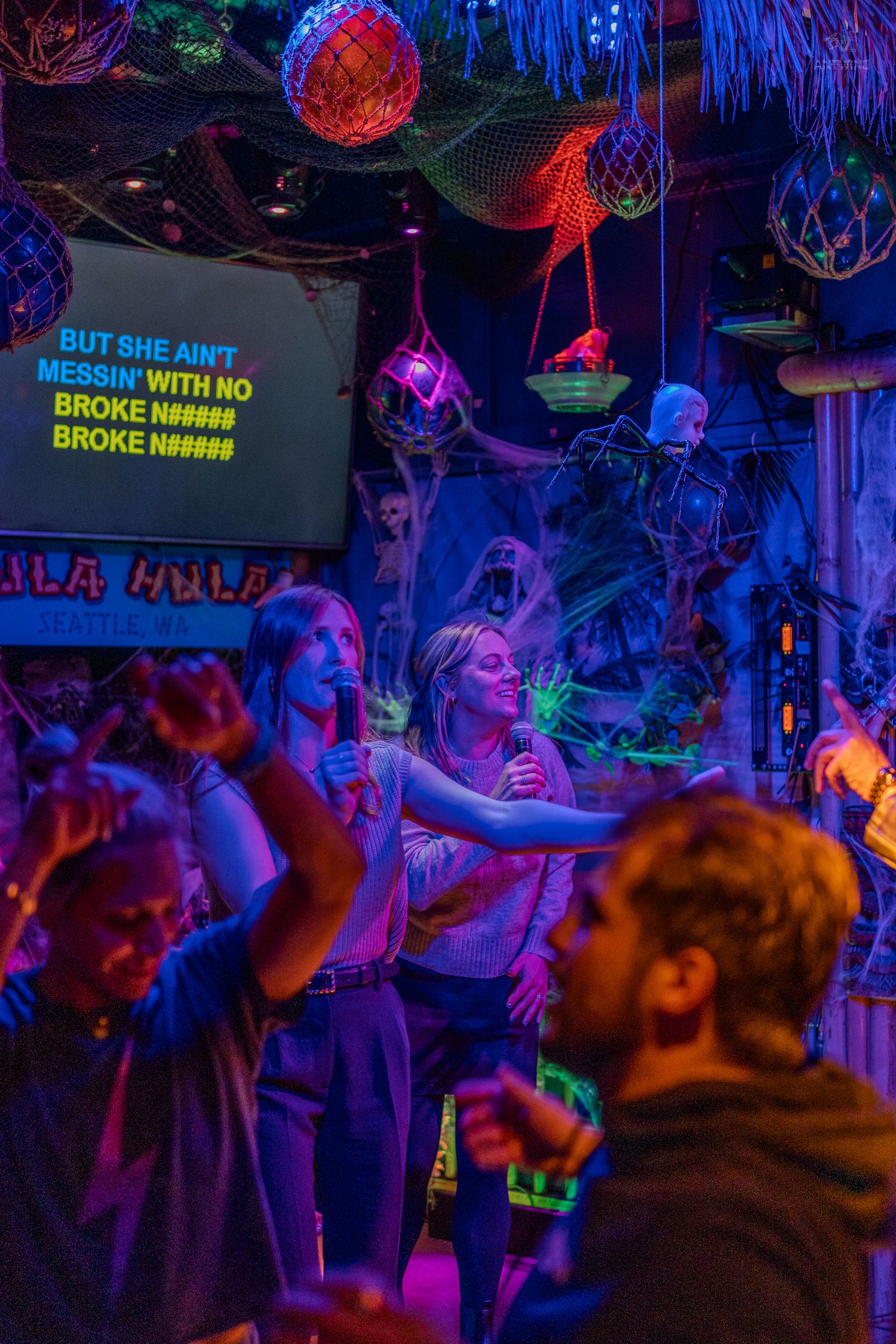 Two women singing karaoke on stage at a neon-lit Halloween tiki bar while guests dance.
