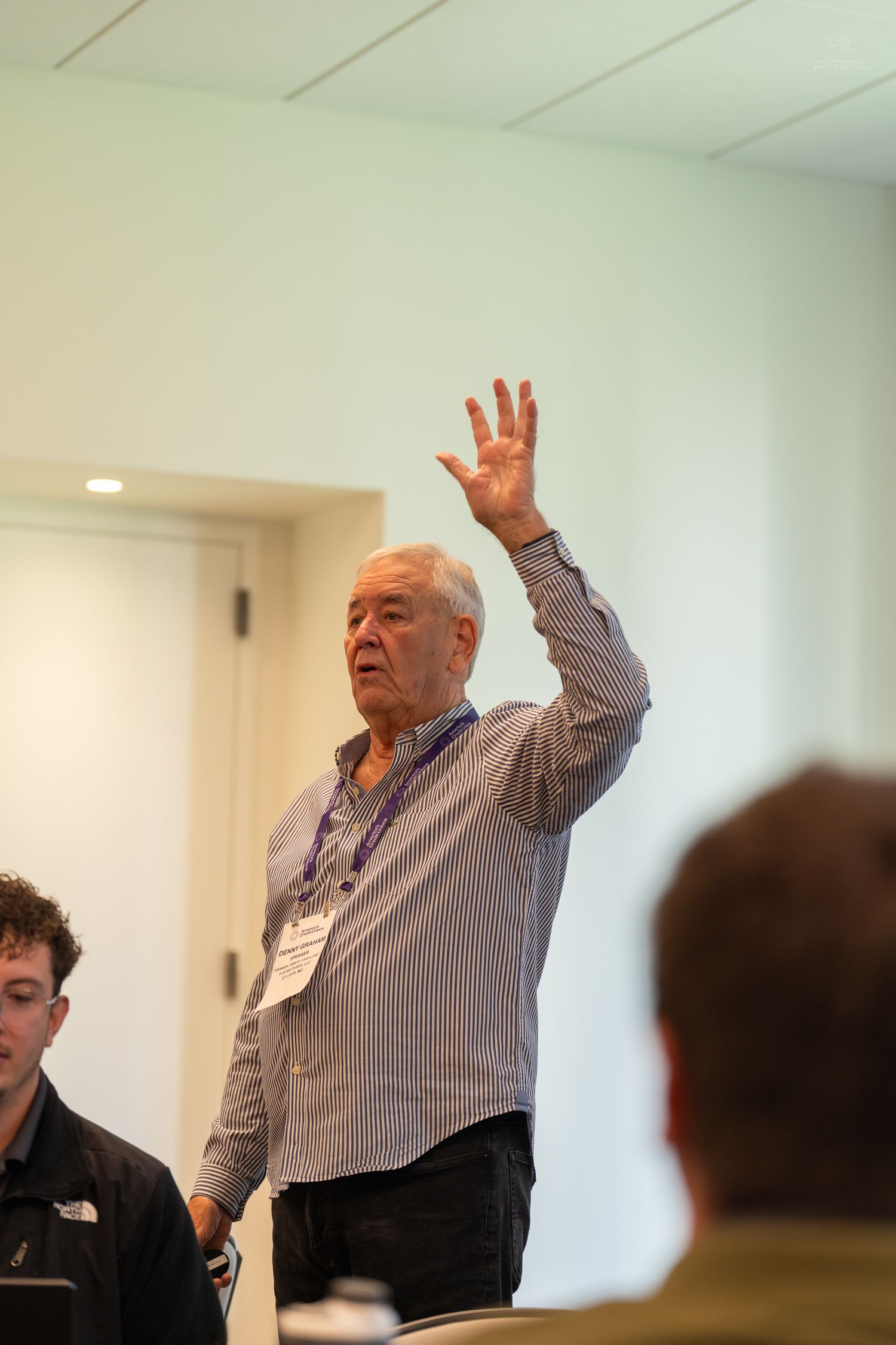 Senior man wearing a name badge raises his hand during an indoor conference or workshop session.