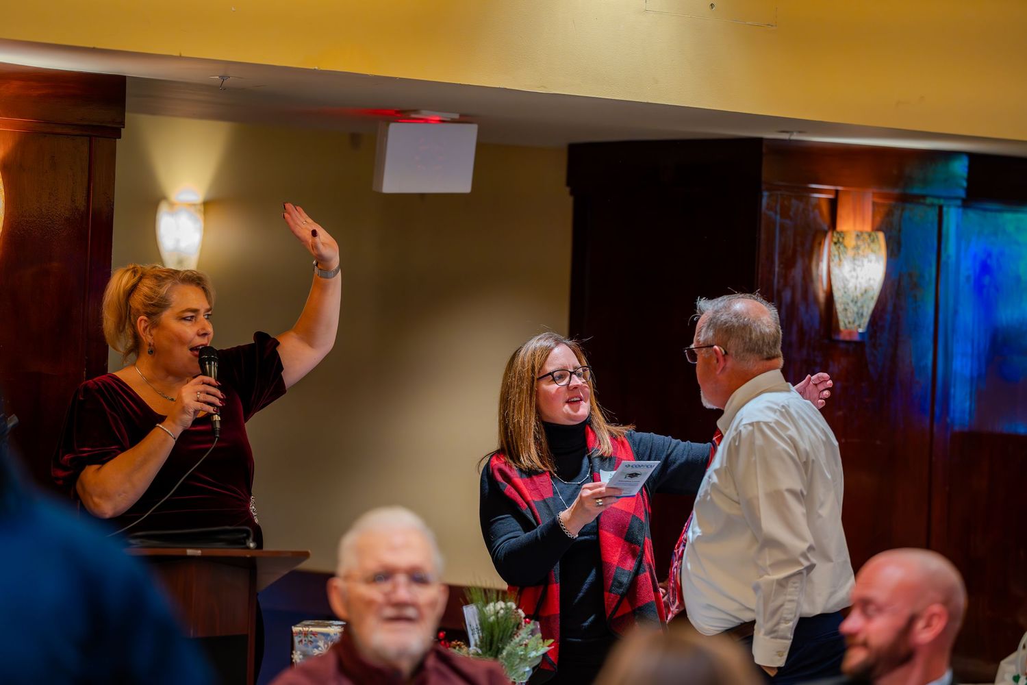 A woman raises her hand during an indoor ceremony while others look on in a warmly lit venue.