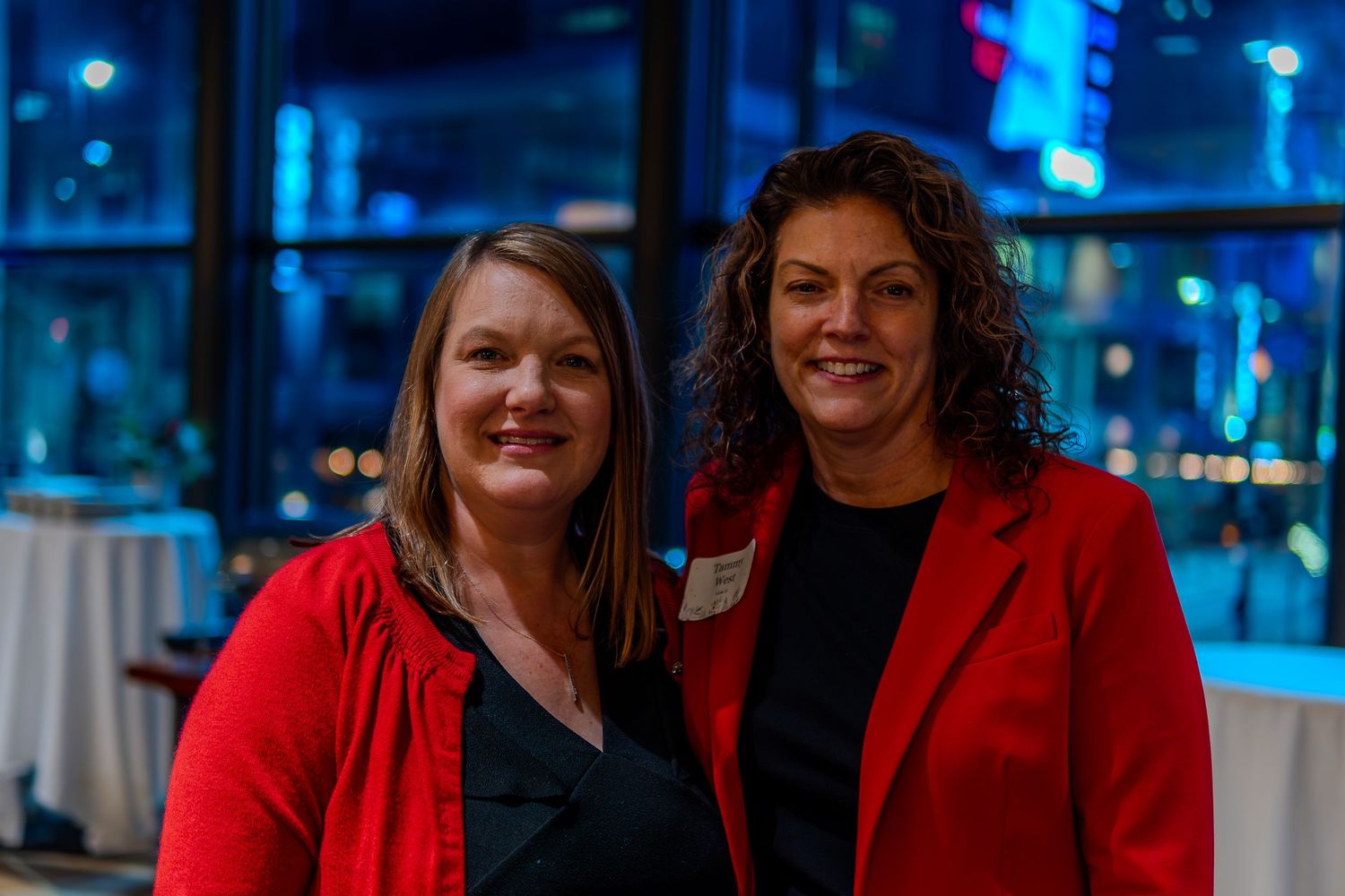 Two women in red jackets smiling together at a formal evening event with city lights in the background.
