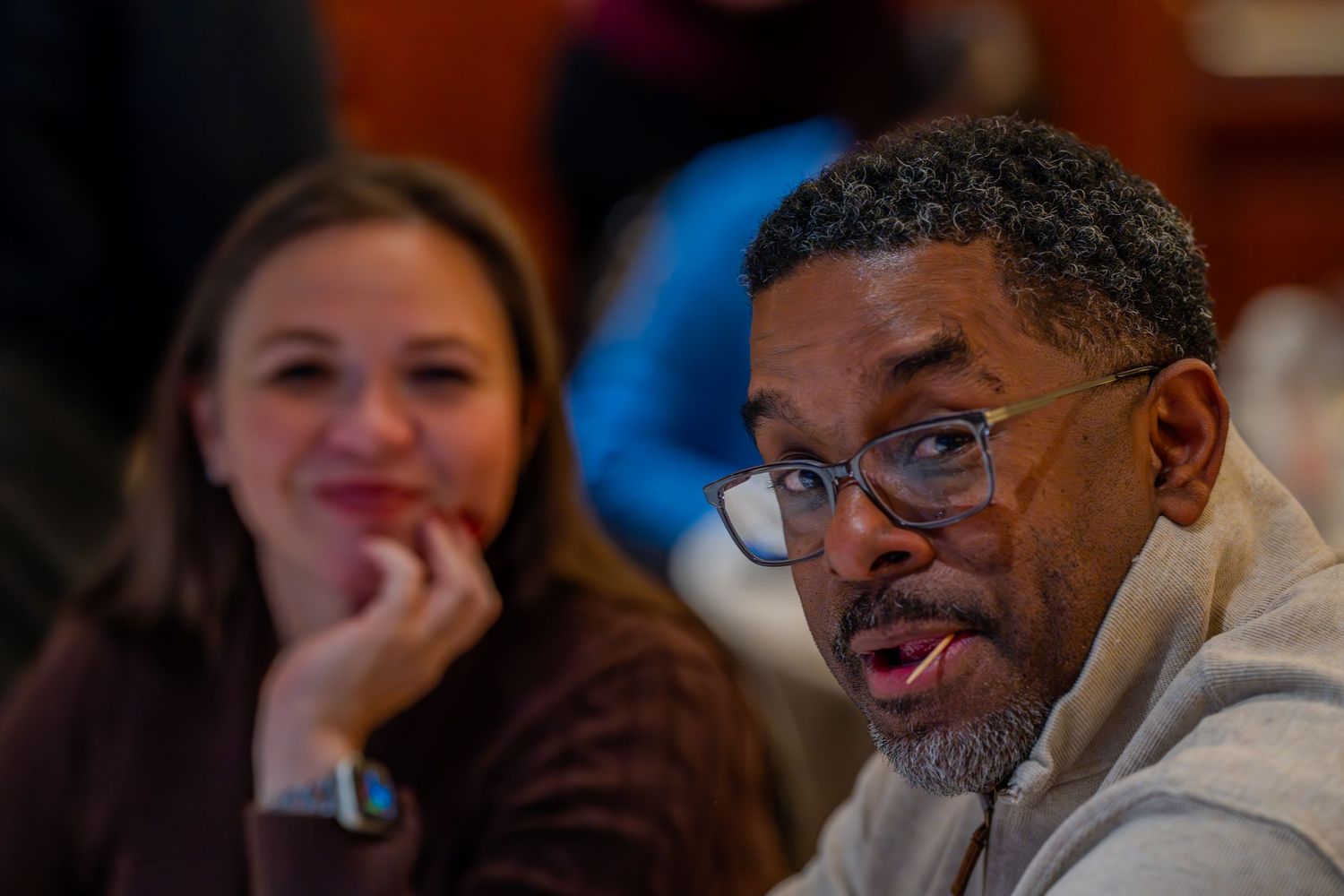 Two people engaged in conversation at an indoor gathering, man with glasses speaking while woman listens attentively.