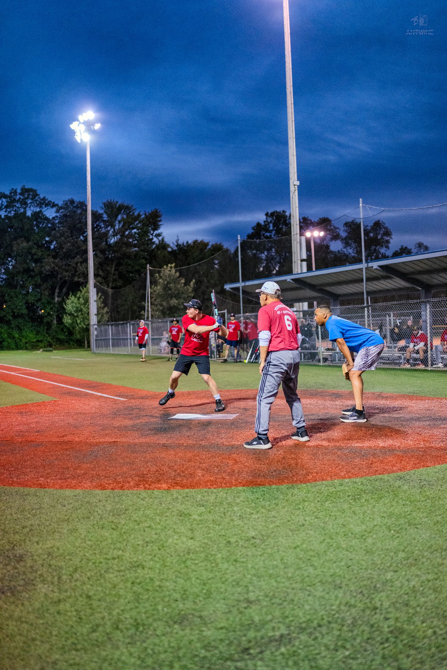 Players compete in a nighttime softball game under bright stadium lights on a red clay baseball diamond.
