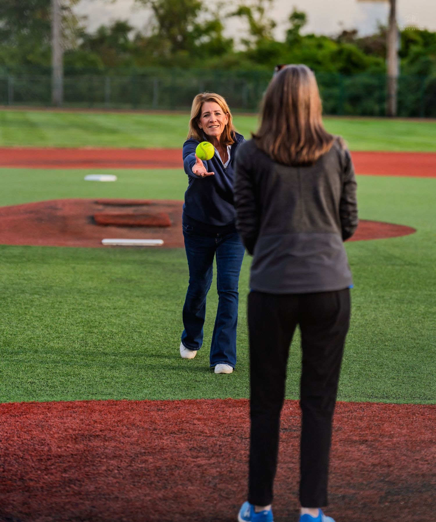 Woman smiling while throwing a yellow softball on a baseball field during evening practice.