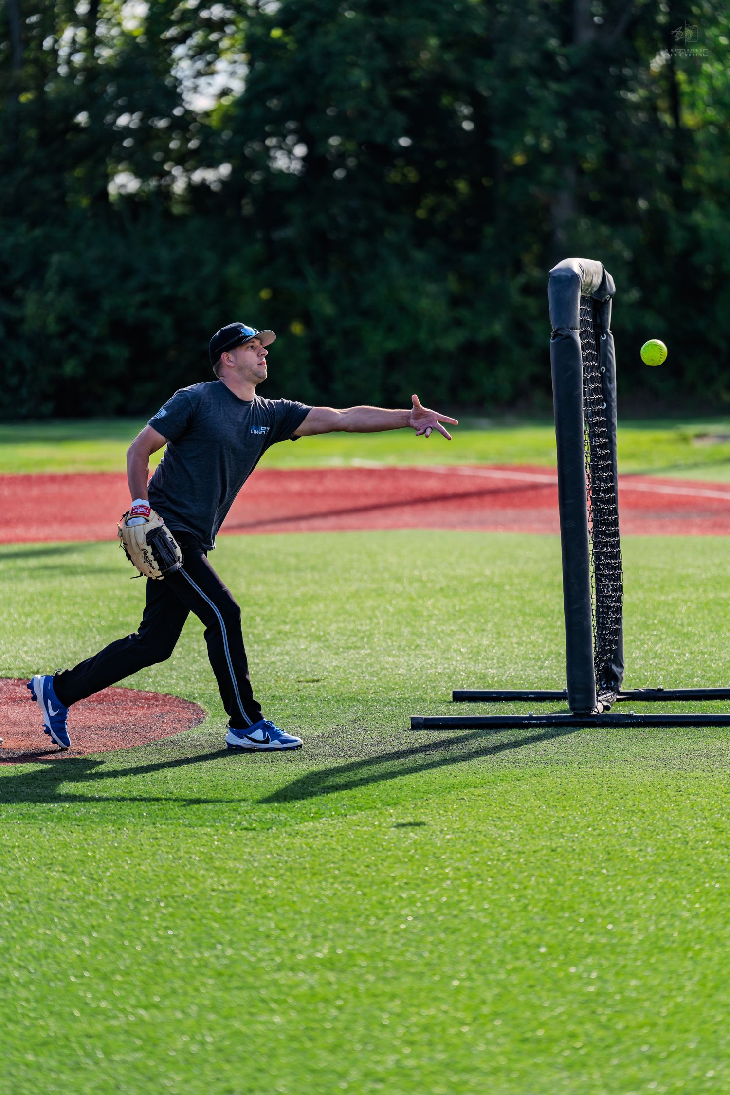 A softball pitcher winds up to throw on a green turf field with a pitching screen during practice.