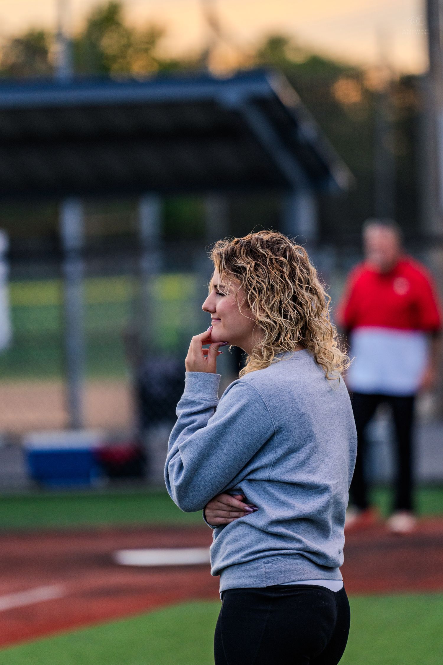A smiling woman with curly blonde hair wearing a gray sweatshirt stands on a baseball field at dusk.