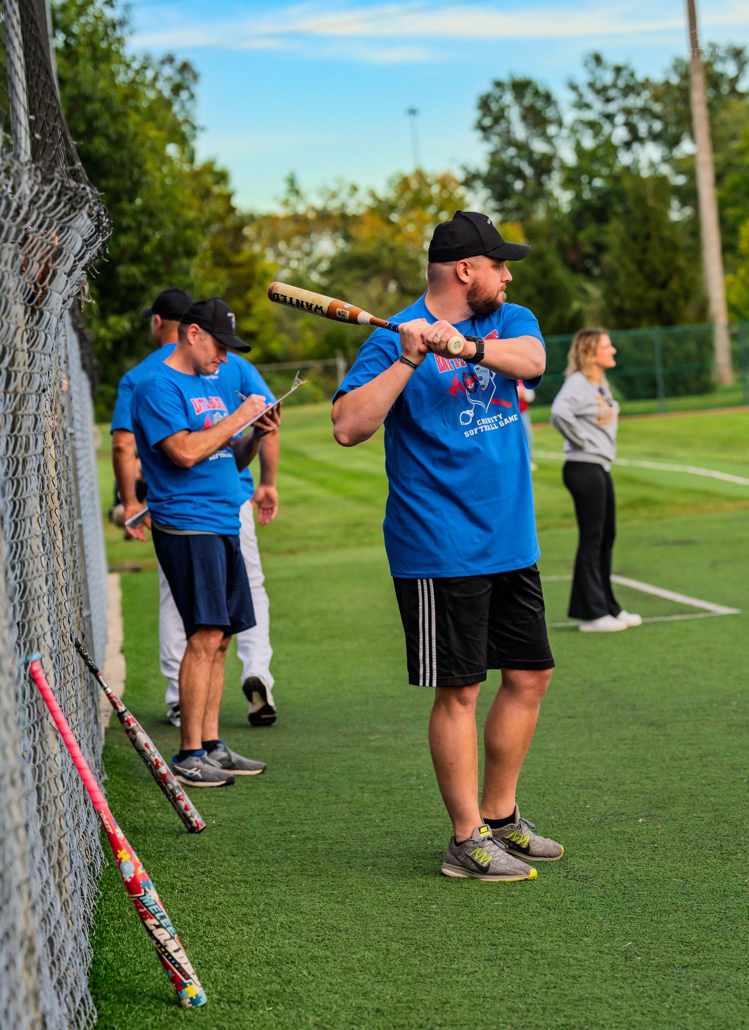 Men in blue shirts practice batting at an outdoor softball field during a recreational league game.