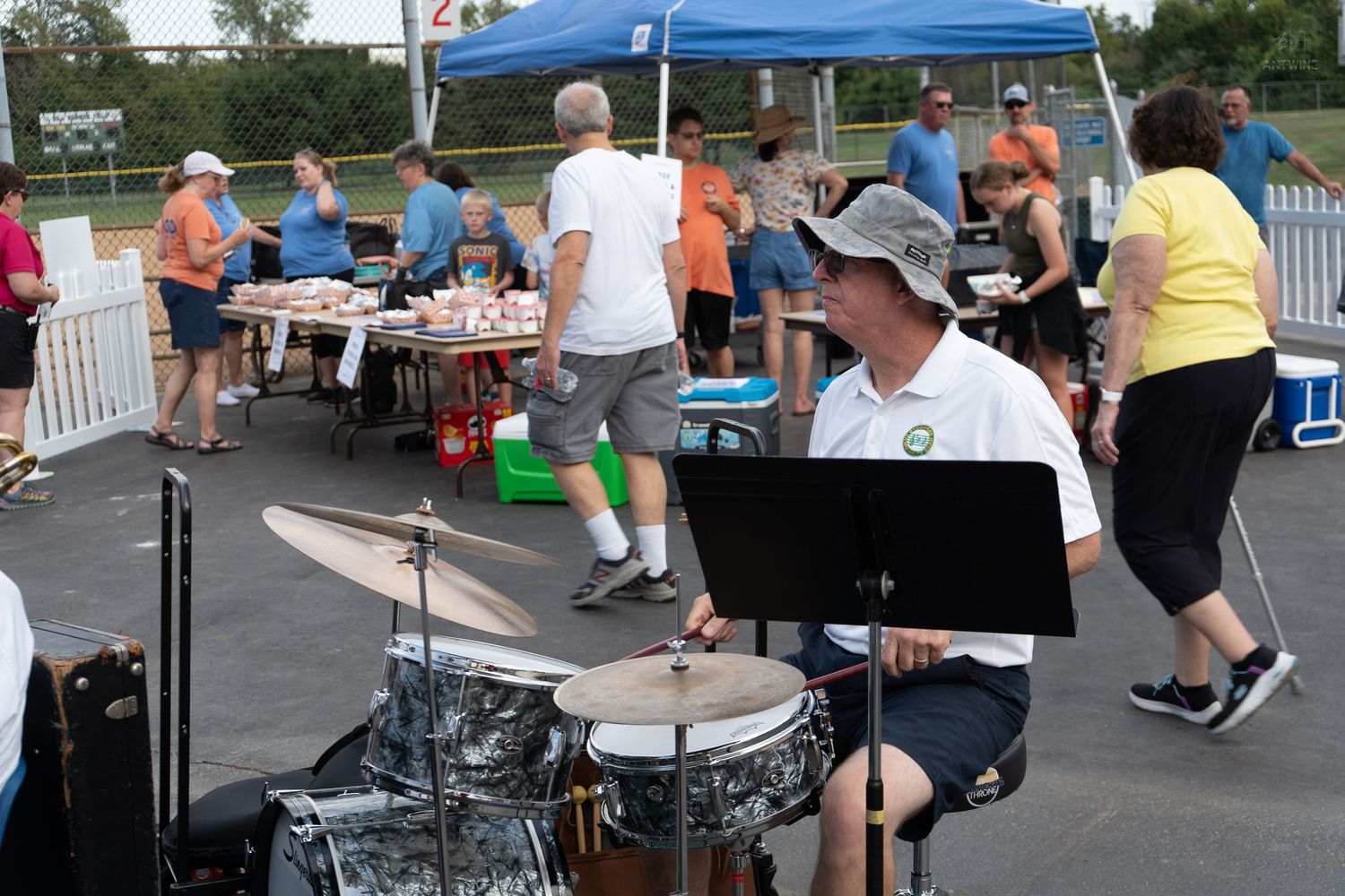 Drummer performs at outdoor community event with people socializing under blue tent on sunny day.