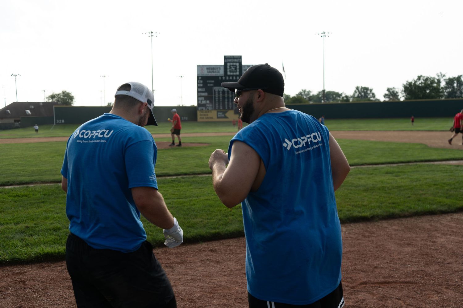 Two OPPCU team members in blue shirts walk on a baseball field during an outdoor event.