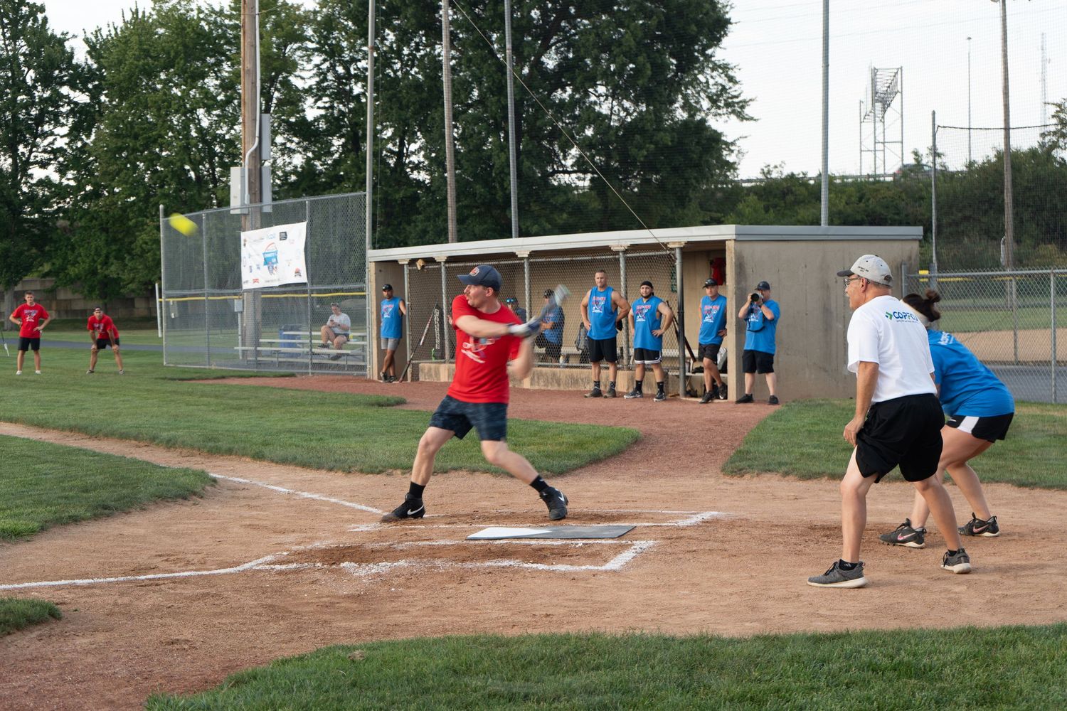 A batter in a red shirt swings at a pitch during a recreational softball game with players and dugout visible.