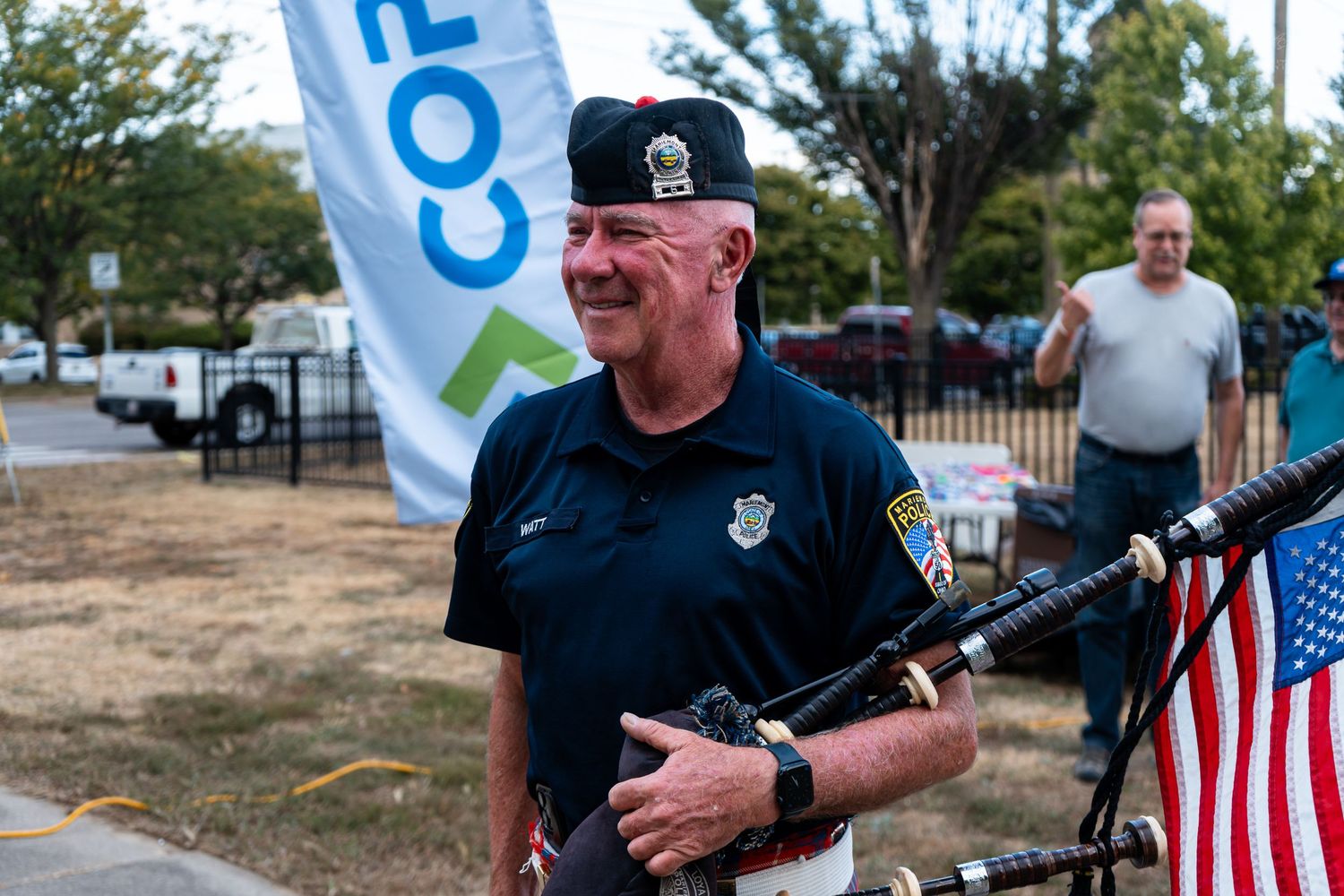 A smiling man in uniform plays bagpipes outdoors near an American flag at a community event.