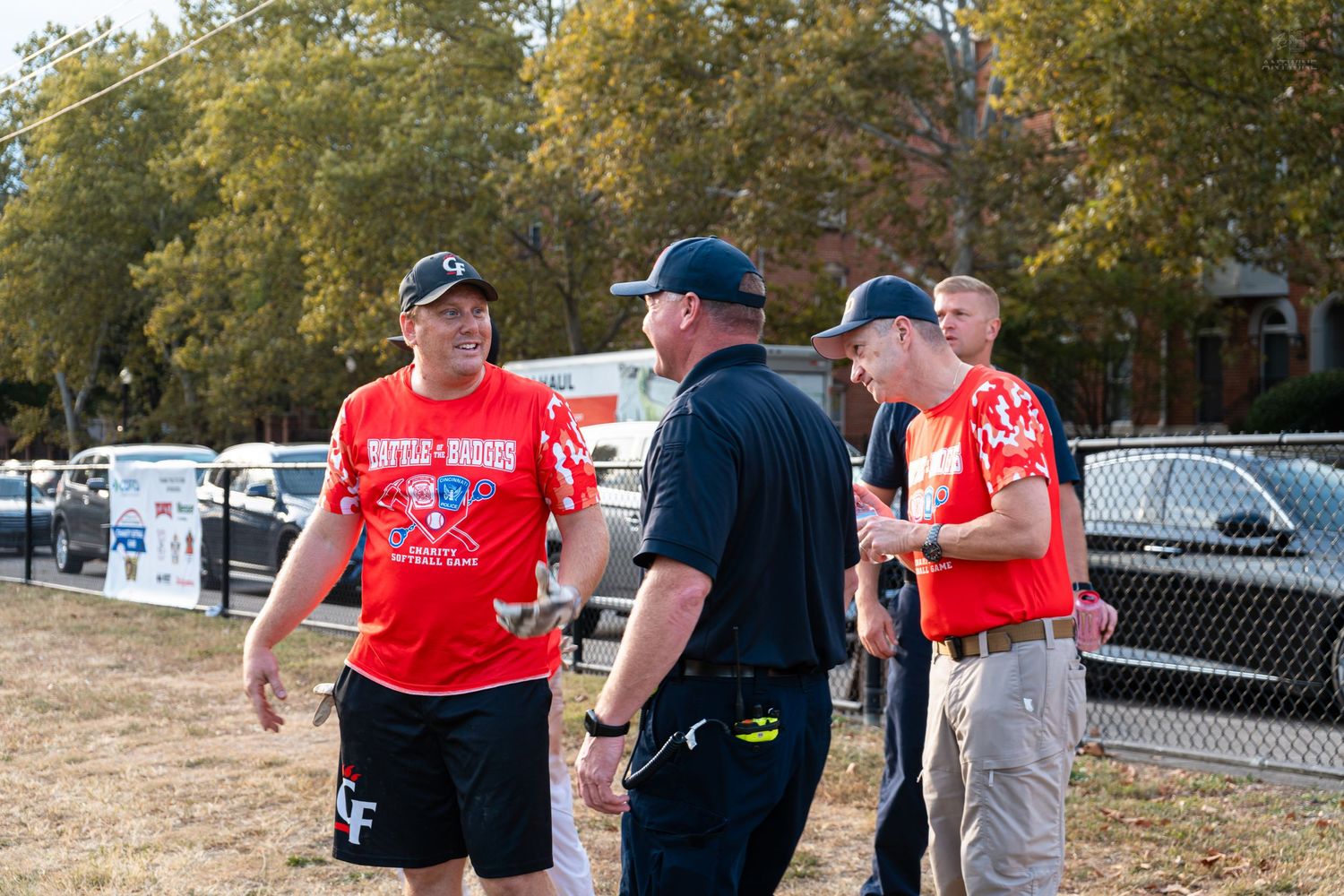 Men in red and black uniforms conversing outdoors at a charity softball game event.