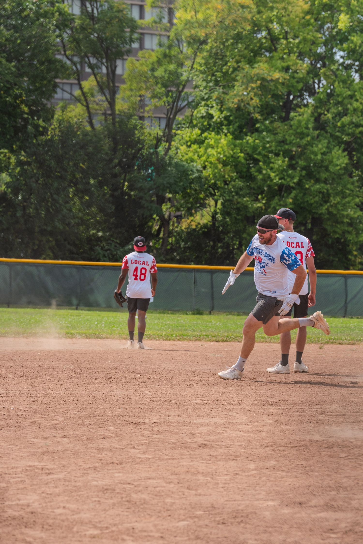Softball players in white Local uniforms on a dirt infield during a sunny recreational game.